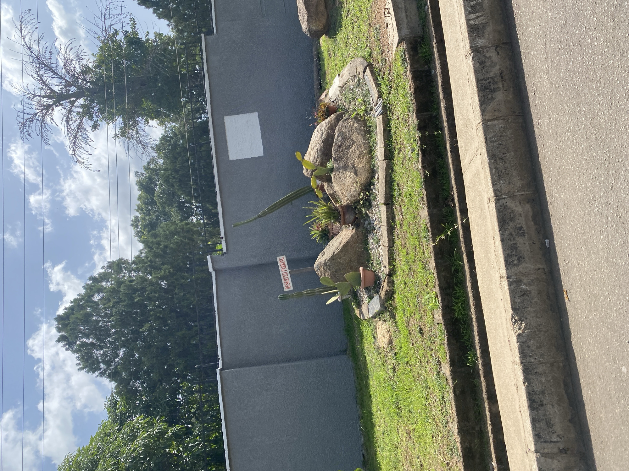 A reserved parking sign is visible in an outdoor scene, likely in Jos, Nigeria, placed near a small rock garden featuring cacti and other succulents. The garden is bordered by a stone curb and is set against a grey concrete wall. Behind the wall, a black metal fence with spikes delineates a property line, beyond which lush green trees and a tall, dry tree with sparse branches are seen under a bright, cloudy sky. The foreground shows a concrete roadside curb and asphalt road.