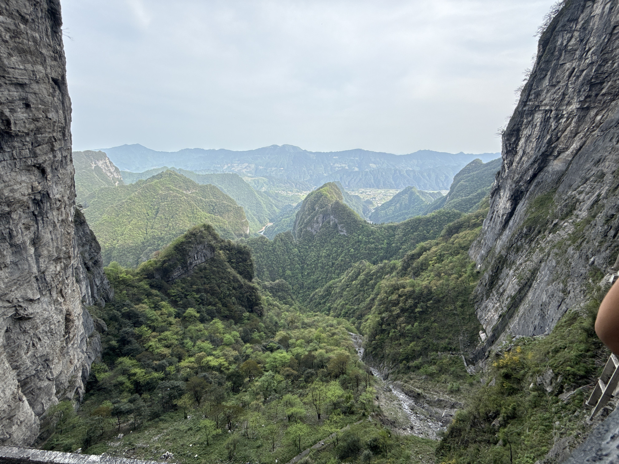 This image captures a sweeping view of the mountainous Zhangjiajie National Forest Park in Yongding District, China. The scene is framed by steep, rocky cliffs on either side, which appear to be part of a gorge or valley. The foreground is dominated by lush green vegetation covering the hillsides and valley floor. A stream or dry riverbed with light-colored rocks is visible winding through the valley. In the distance, a series of layered mountain ranges fade into a hazy, overcast sky. There are no people visible in the image.