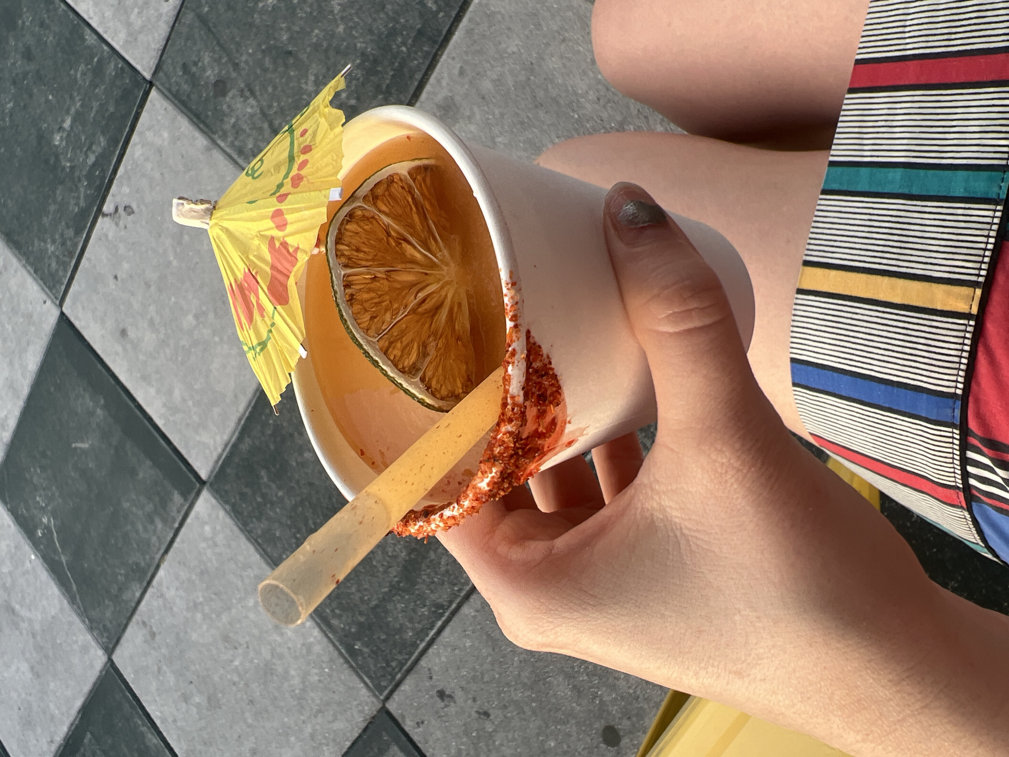 A person is holding a paper cup of an orange-colored beverage with a straw and a dried citrus slice and a small yellow umbrella. The rim of the cup is coated with a red-colored spice. The person's left thumb is visible. The person is wearing a striped garment, which is mostly red, blue, yellow, and turquoise stripes. The background consists of gray and dark gray square tiles. The photo was taken in a city of New York, United States of America.