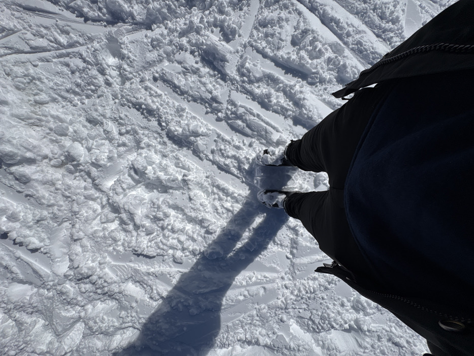 The image depicts a first-person, downward view of a person standing on a snow-covered surface in La Clusaz, France. The subject's dark trousers and dark boots, partially covered in snow, are visible in the lower portion of the frame. A distinct, elongated shadow of a person, including legs, is cast onto the snow to the left of the observer, indicating direct sunlight from the right. The snow surface is extensively disturbed, showing numerous ski tracks and boot prints, suggesting a busy ski slope or snowy trail. The snow is textured with compacted areas and loose, granular piles, indicative of recent activity and varied snow conditions. No other individuals or specific landmarks are visible.