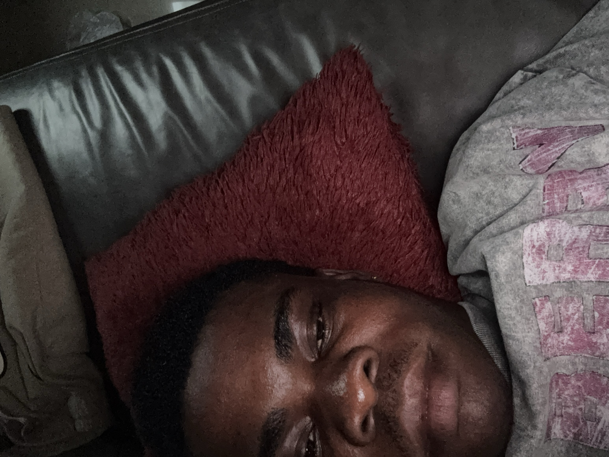 A close-up, low-angle shot shows a Black man lying on what appears to be a dark, possibly leather couch. He is resting his head on a small, red, textured pillow. The man is wearing a gray hooded sweatshirt with a faded pink design. The scene is dimly lit. There is no indication of the image being in Ikot Ekpene, Nigeria.