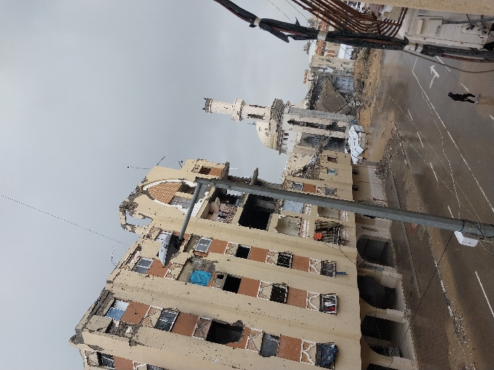 Extensive damage marks a multi-story building and nearby mosque, with shattered windows, crumbling walls, and structural breaches. Debris litters the ground. A solitary figure walks along a rain-slicked street, bordered by partially destroyed structures. Overhead wires hang haphazardly, and a cloudy sky casts a somber light. The scene evokes a sense of devastation and urban decay.