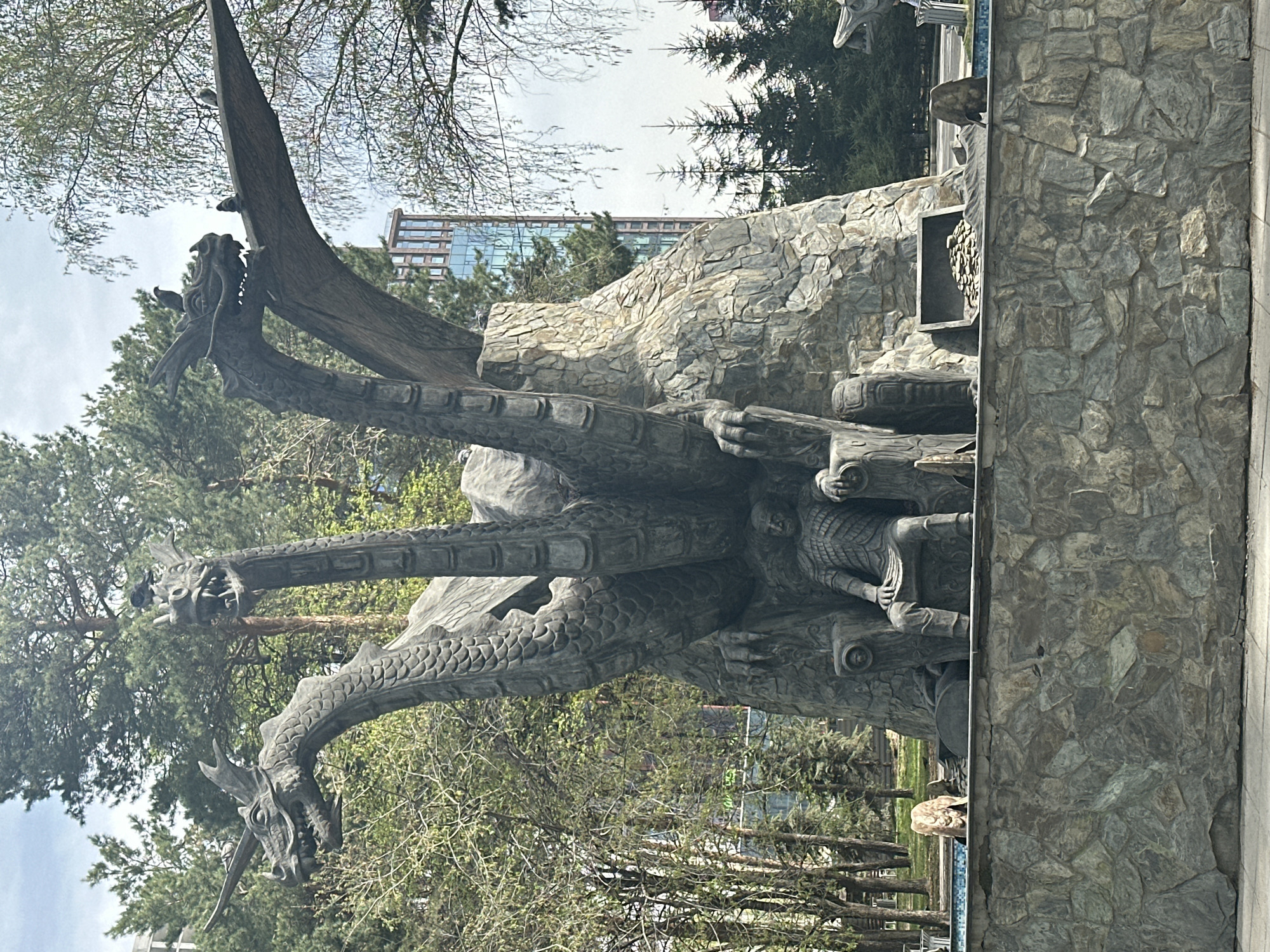 The image depicts a monument of a three-headed dragon in Novosibirsk, Russia. The monument is made of dark-colored stone or metal and features three long, serpentine necks ending in dragon heads. A human figure is sculpted amongst the dragon's coils. The monument appears to be situated in a park or outdoor area, surrounded by trees and foliage. A stone wall or structure is adjacent to the monument, and a modern building can be seen in the background. The sky is overcast.