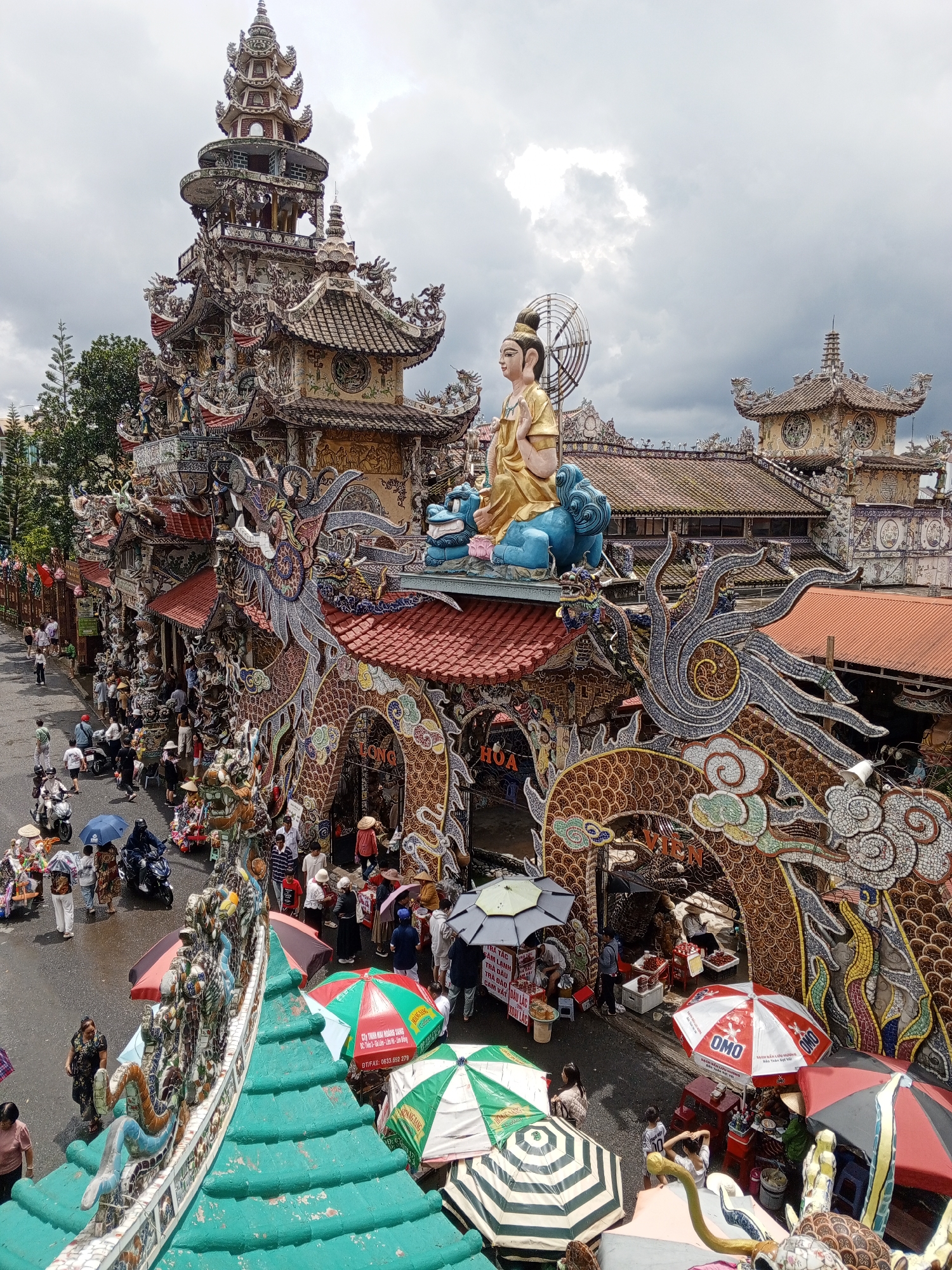 The image depicts a busy street scene in front of a highly ornate temple in Xuân Thành, Vietnam. The temple architecture is characterized by intricate mosaics of ceramic and glass shards, forming elaborate dragon motifs, pagodas, and statues. A prominent golden statue of a seated figure, possibly a deity, is visible atop a blue lotus. The temple entrance is an archway flanked by large, decorative dragons.
The street is populated with many people, some of whom are tourists with umbrellas, suggesting it may be raining or the sun is strong. Several individuals are walking, while others are browsing or interacting with vendors. Motorcycles are present, indicating local transportation. Vendors have set up stalls with red and white umbrellas, selling various goods, possibly food or souvenirs. Colorful market umbrellas are also scattered throughout the scene, providing shade. The ground is wet, suggesting recent rain. The sky is overcast with some brighter areas, indicating a diffused light source. The overall atmosphere is one of a bustling tourist attraction with vibrant, unique architecture.