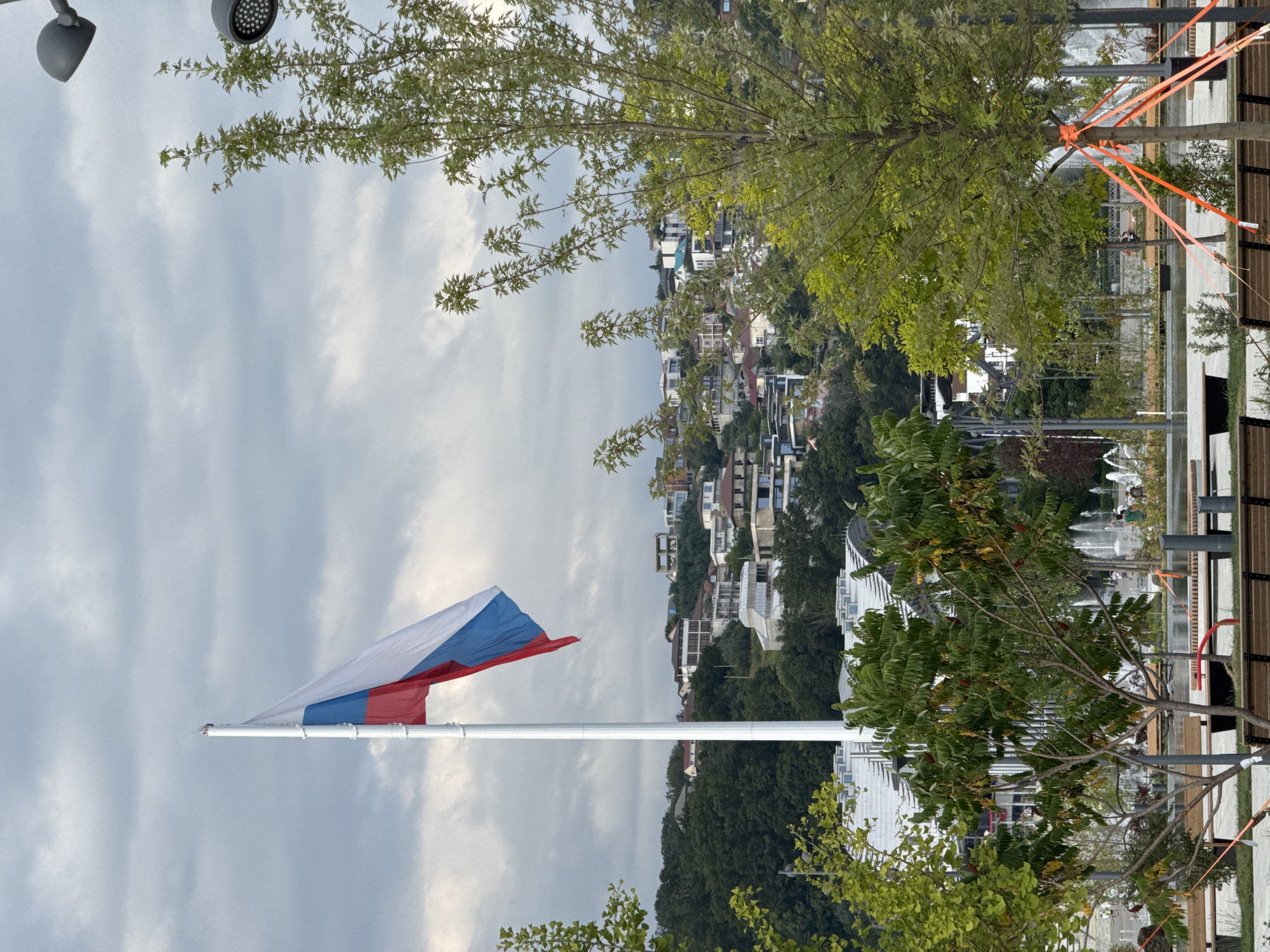 An outdoor, eye-level, vertical shot captures a large Russian flag on a tall white pole in the foreground against a cloudy sky. To the right, a tree with green leaves and orange ribbons tied to its trunk is in the mid-ground. In the background, a hillside is covered with numerous houses and trees. Below the flag, a paved walkway with benches and trees is visible. In the far background, fountains are spraying water. The scene appears to be in a park or public space.