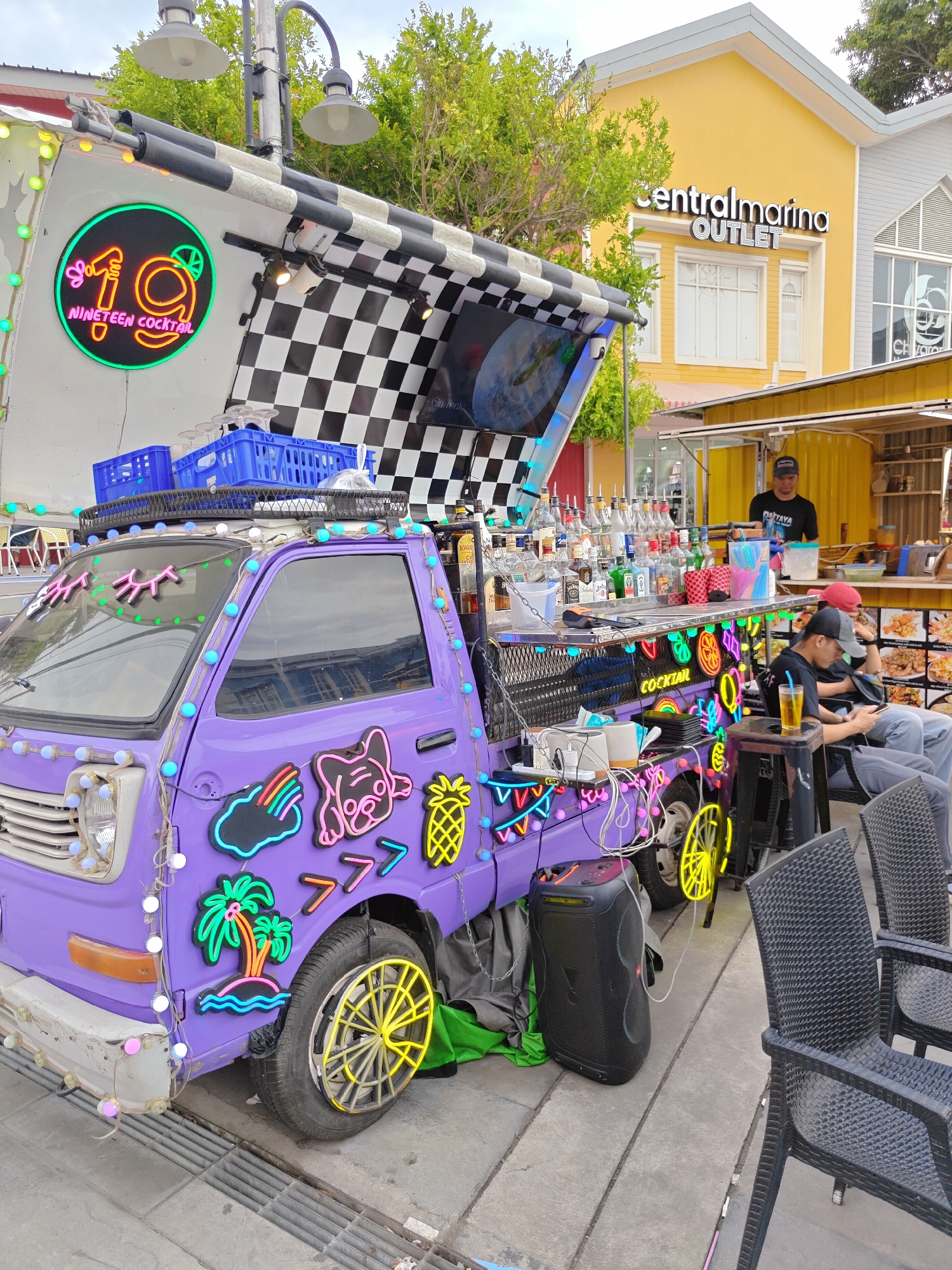 A bright purple, open-air cocktail truck is parked on a paved walkway in Pattaya City, Thailand. The truck is heavily decorated with neon signs, including a large "19 NINETEEN COCKTAIL" sign on the side, as well as neon depictions of eyes, a pineapple, a dog face, a rainbow cloud, and palm trees. String lights in white and pastel colors are also draped around the truck. Behind the truck, a yellow building with "centralmarina OUTLET" signage is visible. In front of the truck, several people are seated at tables. One person, a man wearing a black t-shirt with "PATTAYA" printed on it, is standing behind the counter of a smaller yellow kiosk to the right of the truck. The scene suggests a lively outdoor market or entertainment area.