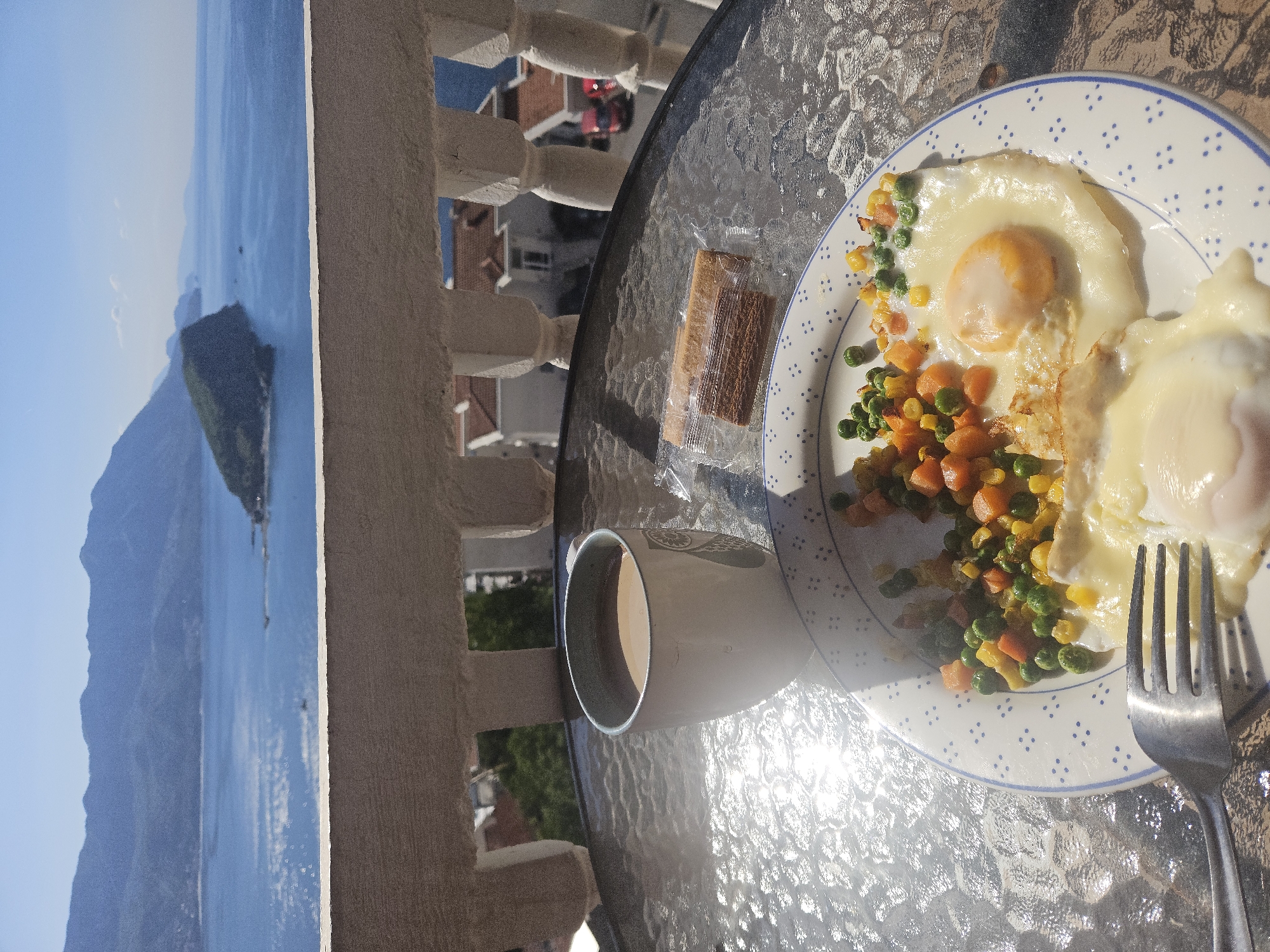 The image shows a breakfast setting on a glass-topped table on a balcony overlooking the Adriatic Sea in Budva, Montenegro. The foreground features a plate with two fried eggs and a side of mixed vegetables (peas, corn, carrots). A packaged cracker sits next to the plate, along with a mug containing a light-colored beverage and a fork. The balcony railing is visible in the background, along with residential buildings and a distant island in the sea. The lighting suggests a sunny morning.
