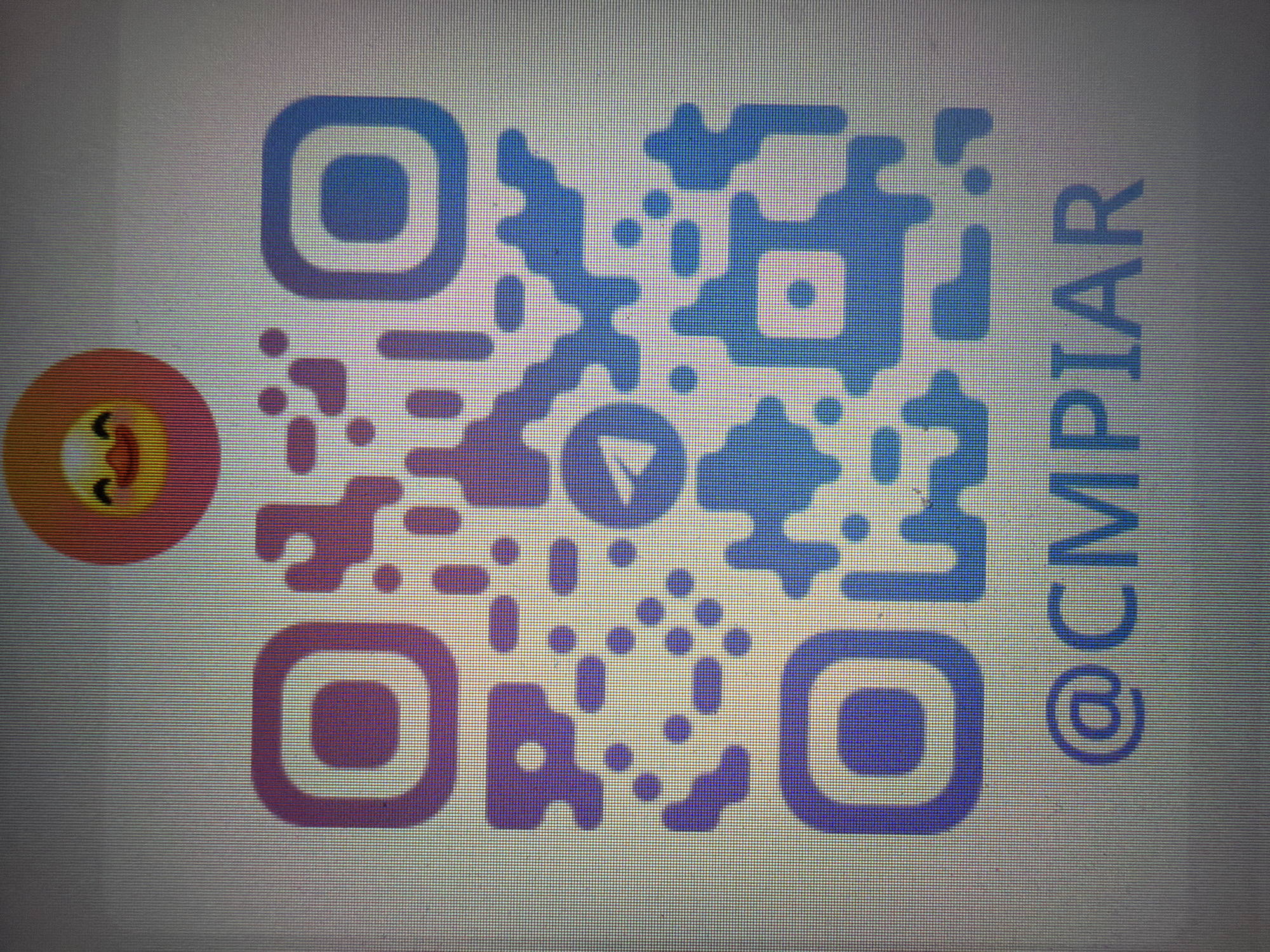 The media file is a close-up, slightly blurry image of a QR code displayed on a screen, likely a monitor or phone. The QR code is predominantly blue and purple, with a gradient effect. To the left of the QR code, there is a circular emoji with a pinkish-orange outline, yellow cheeks, and closed eyes with a smiling expression. On the right side of the QR code, the text "@CMPIAR" is visible in a dark blue, vertically oriented font. The background of the image is a light gray with a visible pixelated grid, suggesting it is a digital display. There is no indication of time of day, weather, or specific location. The overall impression is that of a digital graphic, possibly a social media handle or a link embedded in a QR code.