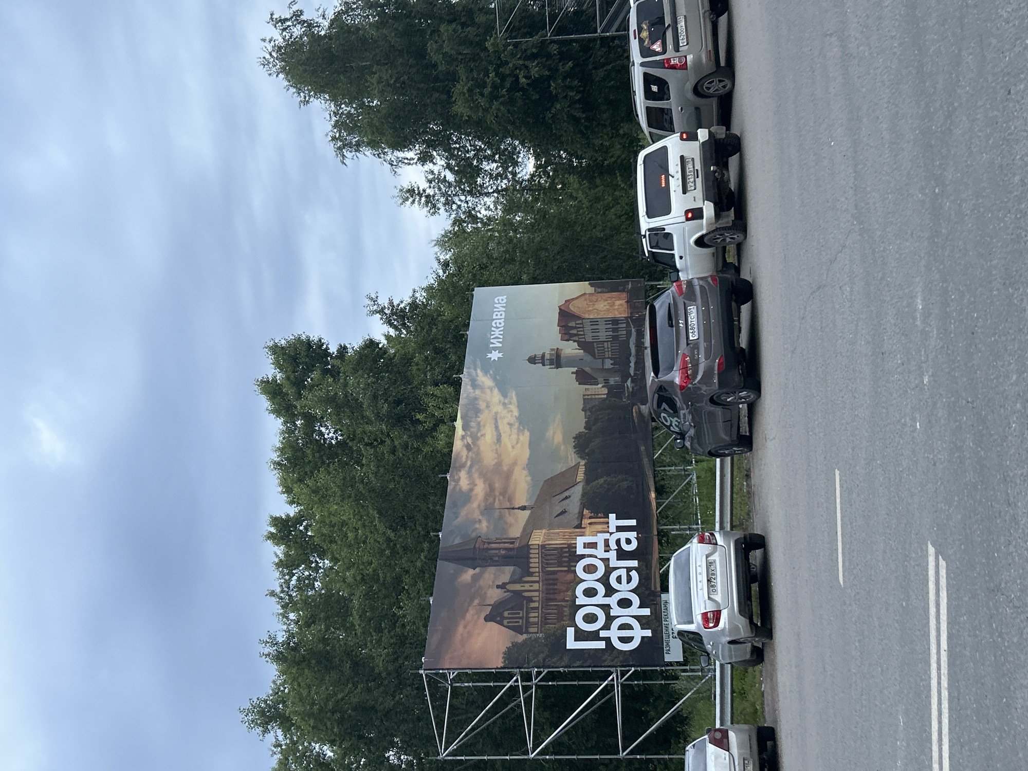 The image shows a billboard in Izhevsk, Russia, advertising a city called "Gorod Freigat" with a picture of a cathedral and a lighthouse. The billboard is supported by a metal scaffolding. In front of the billboard, several cars are parked, including a silver Lada, a white Lada Niva, and a gray Hyundai. The sky is overcast, and the trees in the background are green and lush. The road in the foreground is asphalt with white lane markings. There are no people visible in the image.