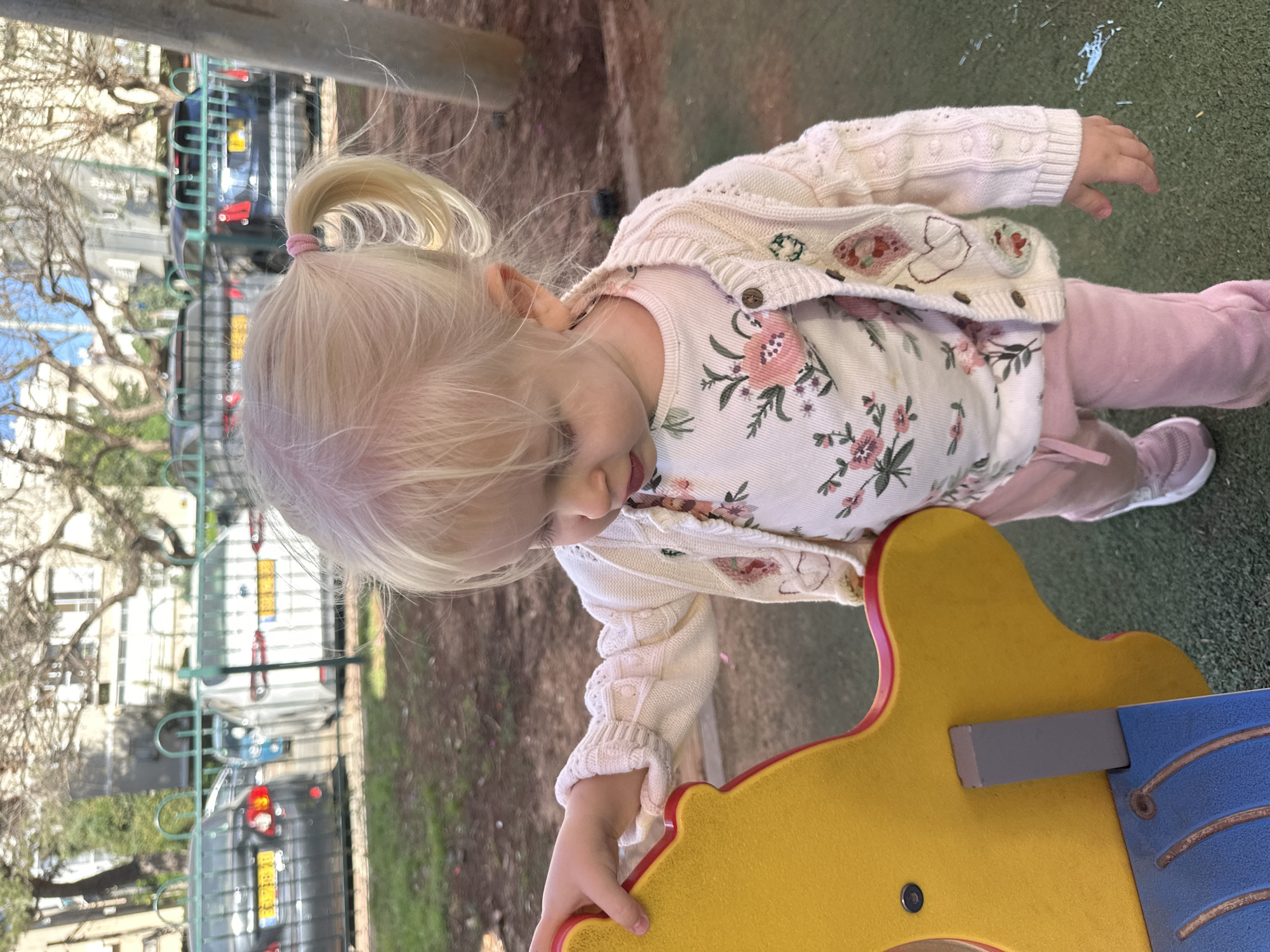 A young Caucasian girl with blonde hair pulled into a ponytail is standing on an outdoor playground. She is wearing a white cardigan with floral patterns, a matching floral shirt, and pink pants. She is holding onto a yellow and blue playground structure. The background shows a cityscape with trees, fences, and vehicles, suggesting an urban environment. Based on the license plates visible on the vehicles, this image was likely taken in Petah Tikva, Israel.