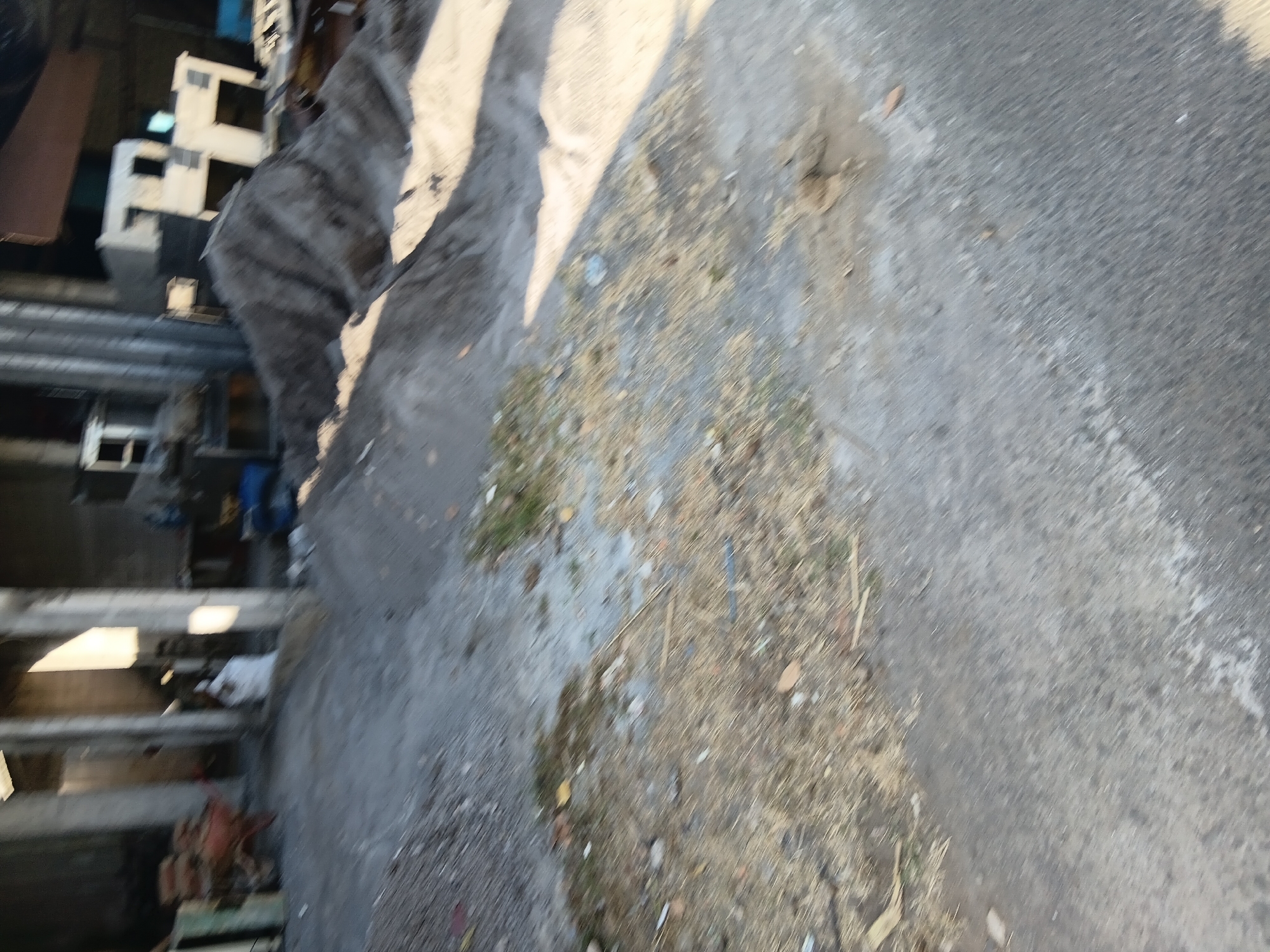 A construction site in Tanggul Wetan, Indonesia, shows a pile of sand and gravel in the foreground. Beyond the sand pile, a partially constructed building with concrete pillars and walls is visible. A wheelbarrow laden with bricks is situated near the entrance of the building. A white bag, possibly containing cement or other construction materials, rests against a pillar. The ground is uneven, with patches of dry grass and scattered debris. The scene suggests ongoing construction activity in a rural or semi-urban setting.
