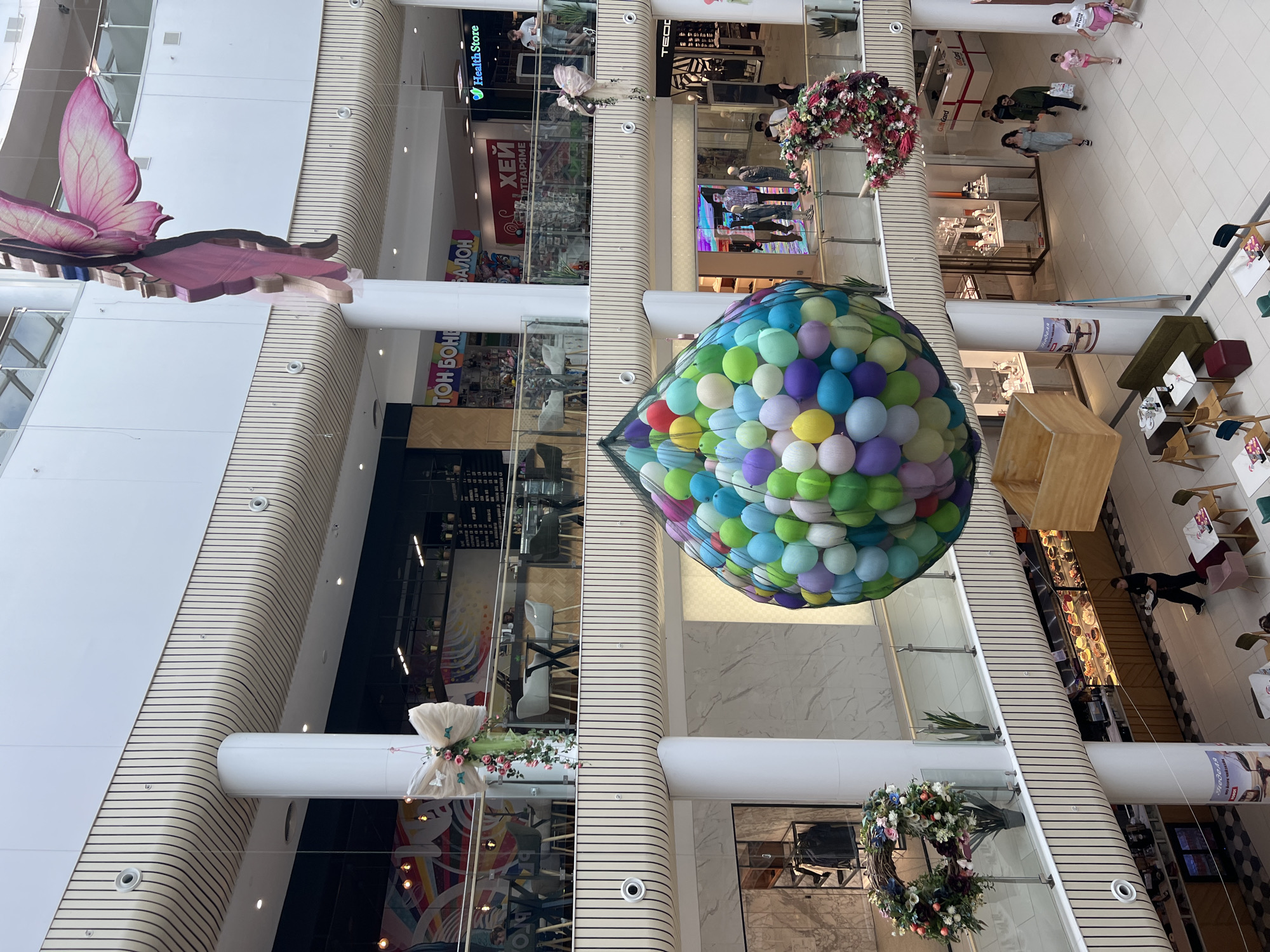 The image depicts the interior of a shopping mall in Plovdiv, Bulgaria. The view is from a high angle, looking down into the multi-story atrium. Several floors are visible, with shops lining the open spaces. The architectural style includes curved walls with linear detailing.
Several decorations are suspended in the atrium: a large, colorful balloon sculpture; a wreath made of flowers; and a butterfly.
People are visible on multiple levels, walking and interacting. Shops can be seen, with various displays of merchandise. Some shops appear to be cafes or restaurants. Furniture, like tables and chairs, are visible in some areas. The floor is tiled in a light color. Overall, the environment is brightly lit and modern.