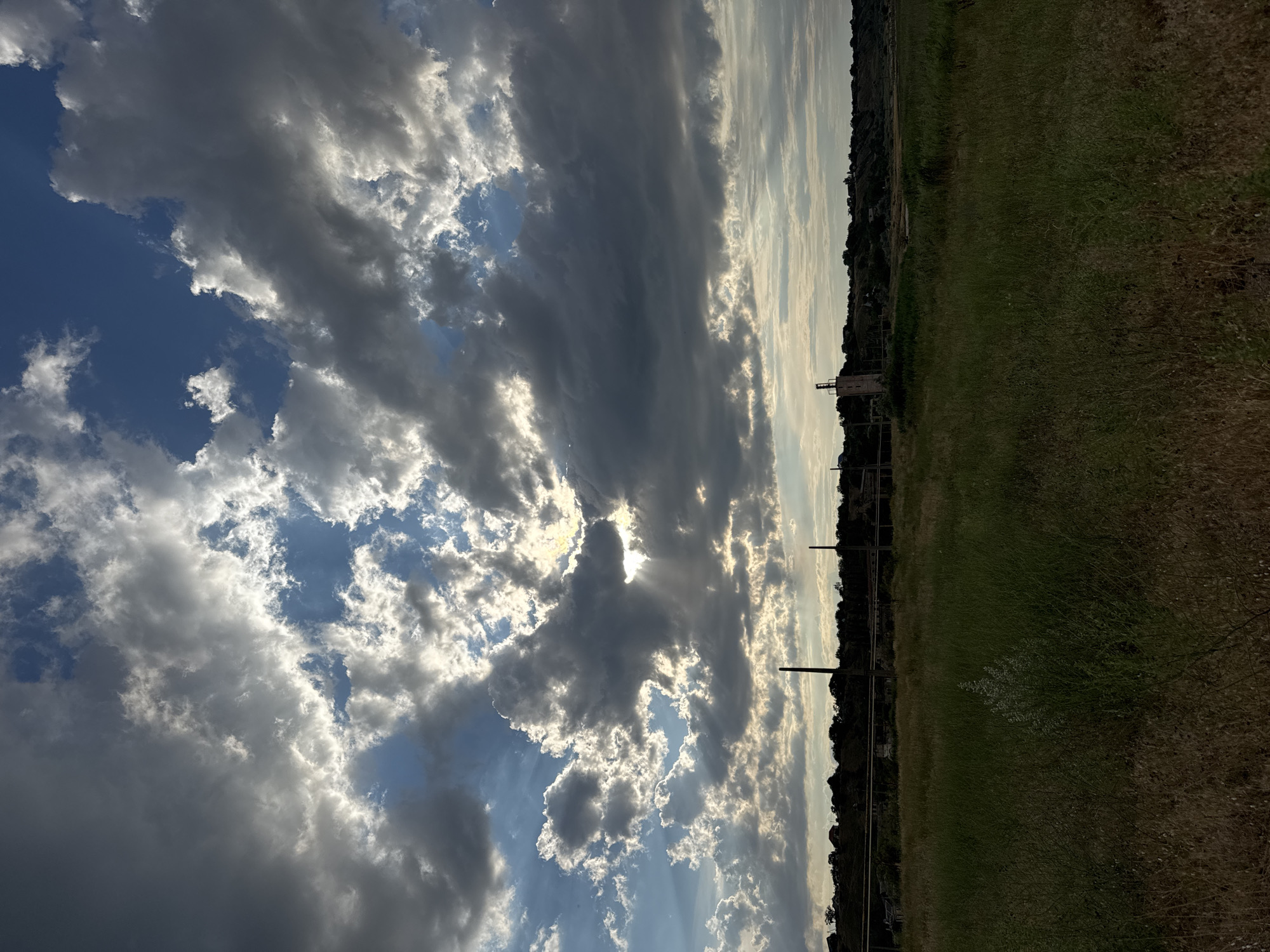 This is a vertical, outdoor, landscape photograph of a cloudy sky over a field with some industrial structures in the distance. The sky dominates the left side of the frame, filled with a mix of dark gray and bright white clouds, with patches of blue sky visible. The sun is partially obscured by the clouds, creating rays of light that pierce through. The right side of the frame shows a grassy field that transitions to a line of trees and distant buildings, with what appear to be industrial towers or smokestacks. The lighting suggests it is late afternoon or early evening. The overall mood is somewhat dramatic due to the contrast between the dark clouds and the bright sunlight. There is no visible text in the image.