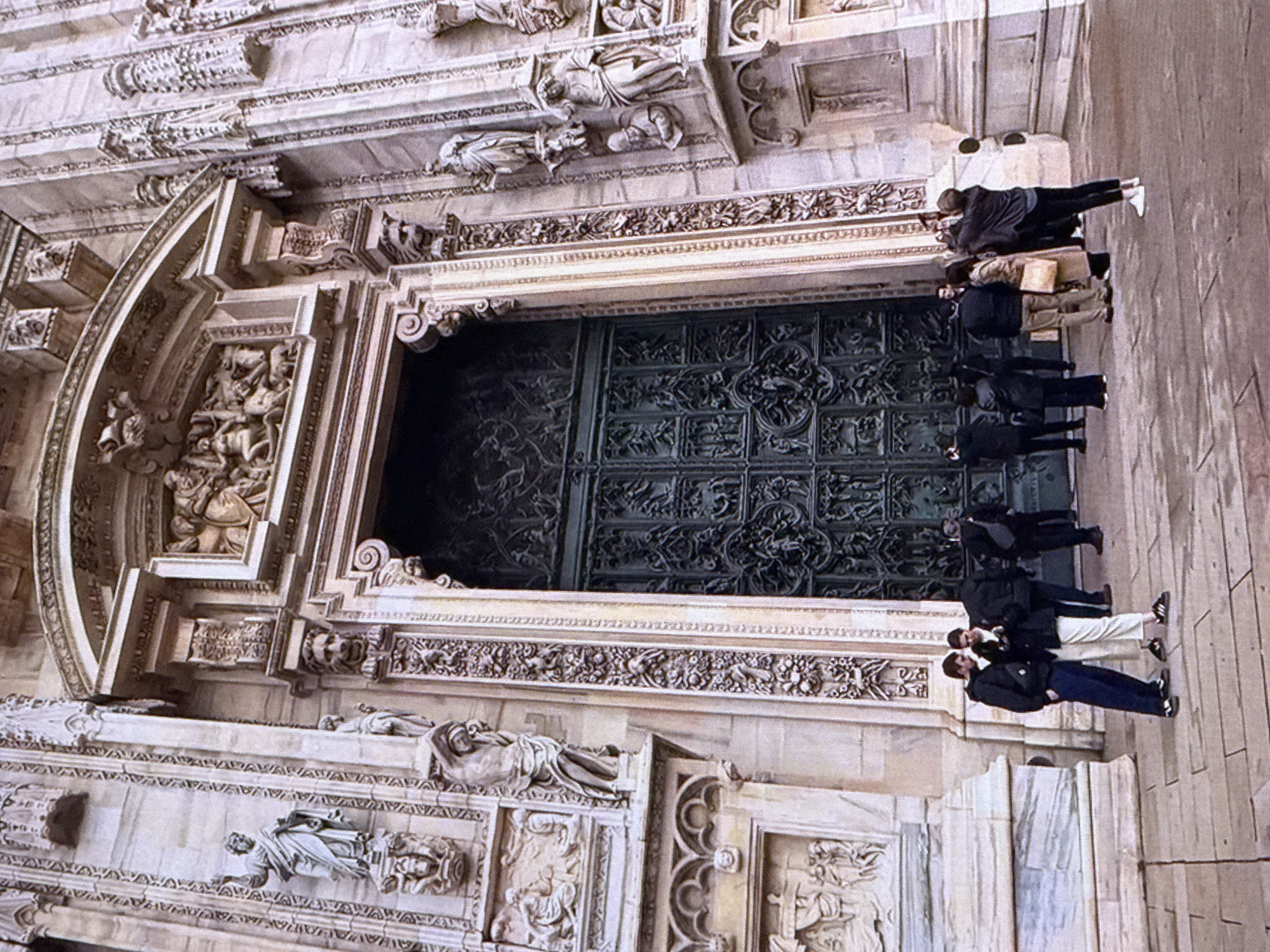 The image shows a group of people standing on the paved ground in front of a large, ornate building. The building features a grand entrance with intricate carvings and large, dark double doors. Several statues and detailed architectural elements adorn the facade. The people are casually dressed and appear to be tourists, some are looking at their phones, while others are taking pictures. The lighting suggests it is daytime, and the overall scene depicts a popular tourist site. The location of the building is not Dubai, United Arab Emirates, but rather a European cathedral, likely the Duomo di Milano in Milan, Italy, given the distinctive Gothic architecture and the detailed sculptures.