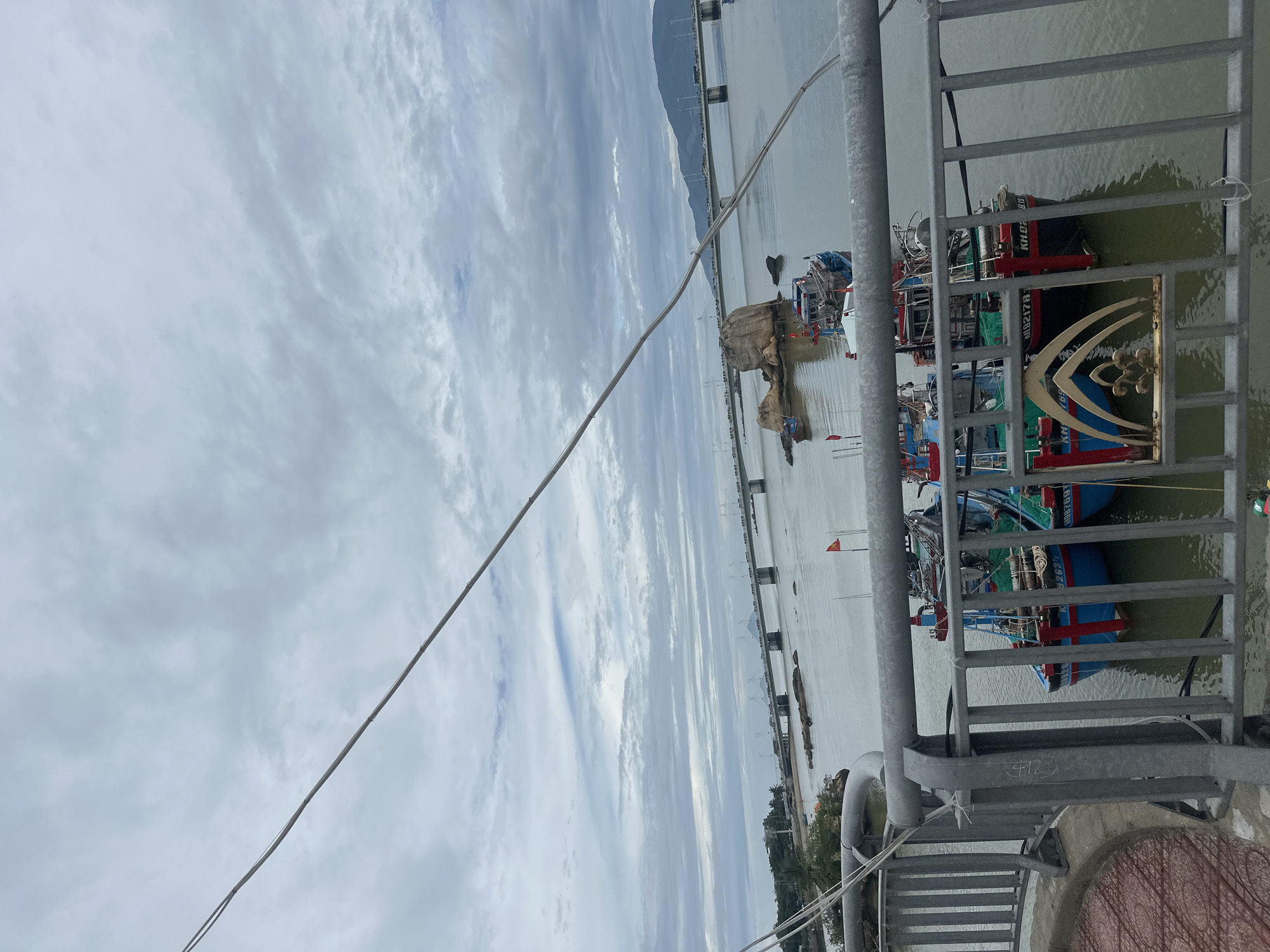 The image shows a view from a bridge in Bac Nha Trang Ward, Vietnam, looking out over a body of water with numerous fishing boats docked. The sky is overcast with a significant amount of cloud cover. The bridge has metal railings visible in the foreground, with an ornate, decorated section integrated into the railing. In the distance, a long bridge stretches over the water toward the land, connecting to a mountainous region where wind turbines are visible.