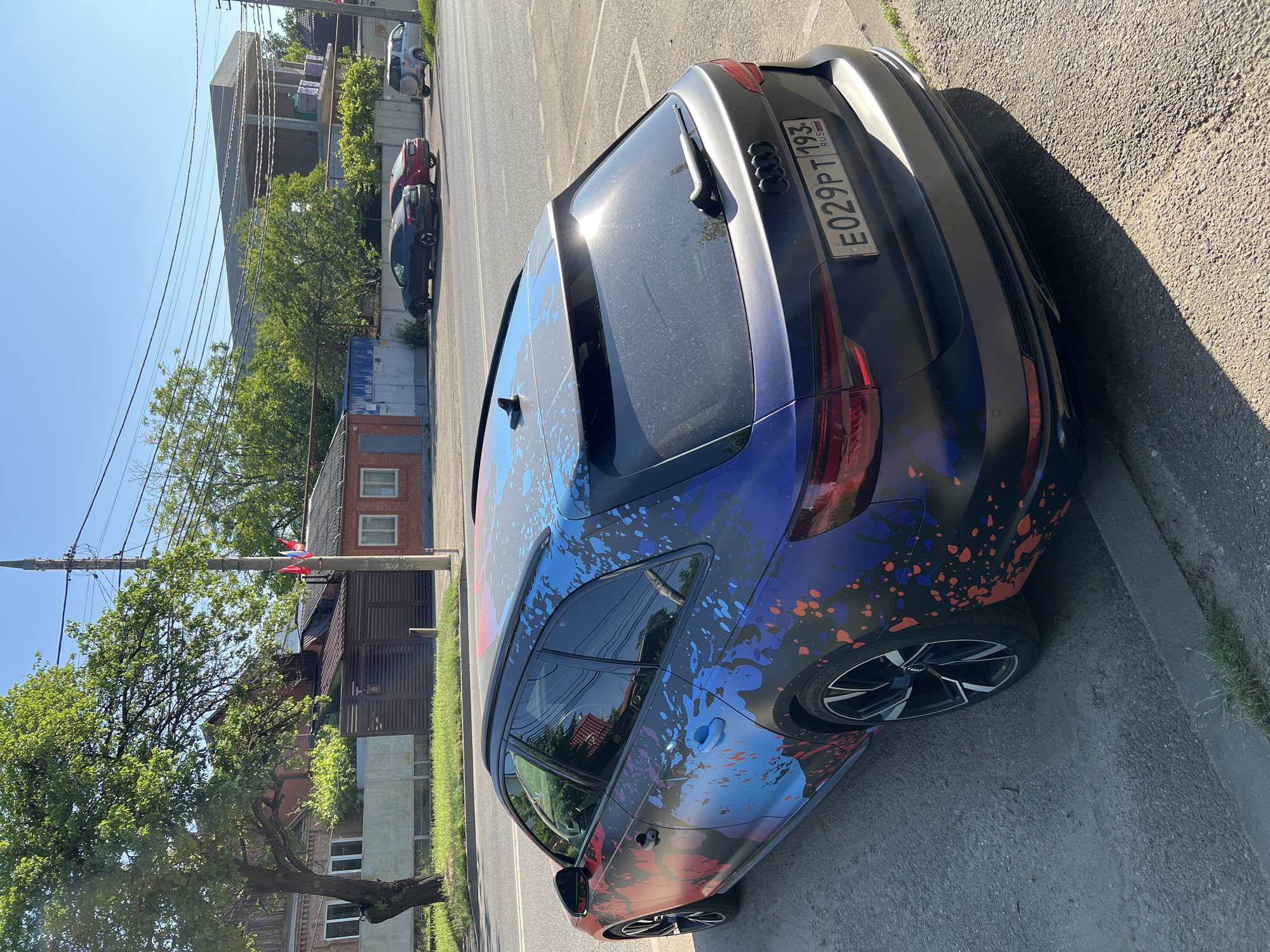 This image captures a street scene in Krasnodar, Russia, featuring a uniquely customized Audi A4 Avant (B9 generation) parked on a dirt and gravel shoulder. The vehicle, observed from a rear-right perspective, features a matte dark grey or black base wrap adorned with an abstract, vibrant paint splatter design in shades of blue, purple, orange, and red, covering its side and roof. Its rear license plate, "E029PT 193 RUS," confirms its registration in Krasnodar Krai. The car is fitted with black multi-spoke alloy wheels with silver accents.
Across a paved asphalt road, several structures are visible. These include older, brick-built residential-style buildings, some with fences and private gates. Further down the road, a larger, unfinished concrete structure with a distinctive cantilevered roof design stands out, suggesting ongoing construction. Adjacent to the road, a utility pole displays three small flags; one is clearly the Russian national flag (white, blue, red horizontal stripes), with two other smaller flags, one predominantly red, directly below it.
The environment is characterized by abundant green foliage, including mature trees with dense canopies and various bushes. Overhead, a network of utility lines spans across the street. Three other vehicles are parked further along the opposite side of the road: a black sedan (possibly an Audi A5 Sportback), a red sedan (resembling a Mazda 6), and a silver SUV (possibly a Lada Niva Travel). The scene is illuminated by bright, direct sunlight, casting distinct shadows from the parked Audi onto the ground, indicating a clear, sunny day. No individuals are present in the frame.