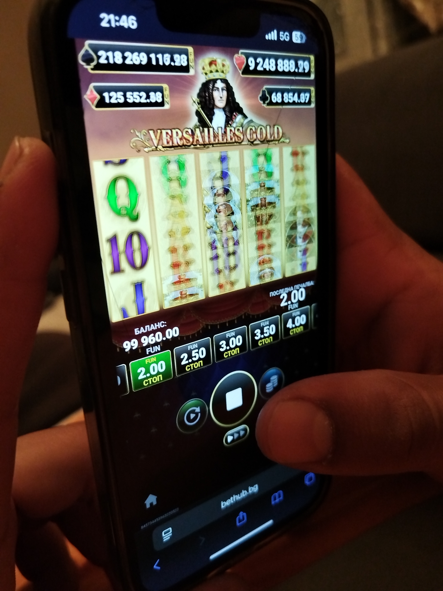 A person is holding a smartphone displaying a virtual slot machine game called "Versailles Gold." The phone's screen displays various game elements: a playing field with spinning reels, digital balances represented by card suits, and betting options. The phone displays 21:46 on the top left corner and 5G at the top right. The phone's browser is open at the bottom of the screen, with "bethub.bg" displayed in the address bar. The background is blurred and does not provide any additional environmental context.