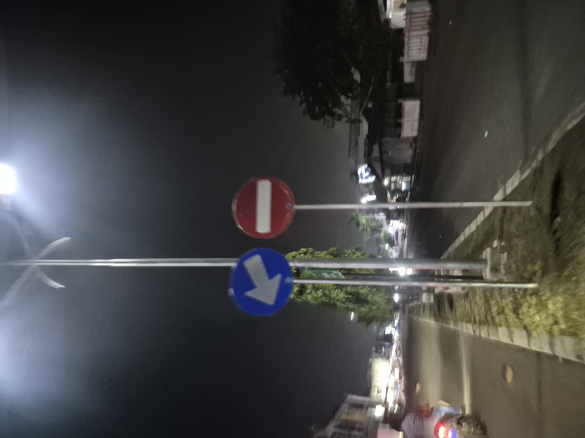 This nighttime image, captured in Lhokseumawe, Indonesia, presents a street scene dominated by two traffic signs. The sky is dark, characteristic of night, with artificial lights creating bright spots and glare, particularly a strong light source on the left side of the frame.
In the center, two circular traffic signs are mounted on a pole. The upper sign is a red circle with a horizontal white bar, universally recognized as a "No Entry" or "Do Not Enter" symbol. Below it, a blue circular sign displays a white arrow pointing downwards, typically indicating a mandatory direction such as "Keep Right" or specifying a lane.
To the right of the signs, a paved road is visible, bordered by a curb and a strip of ground covered with sparse vegetation. In the background, dimly lit buildings and trees line the street, indicating an urban or semi-urban setting. The road surface appears reflective in areas, possibly due to wet conditions or the glare from streetlights. In the bottom right corner, a blurry red light suggests the taillight of a moving vehicle, likely a motorcycle or scooter, hinting at ongoing traffic activity. No discernible text is visible other than the symbols on the traffic signs.