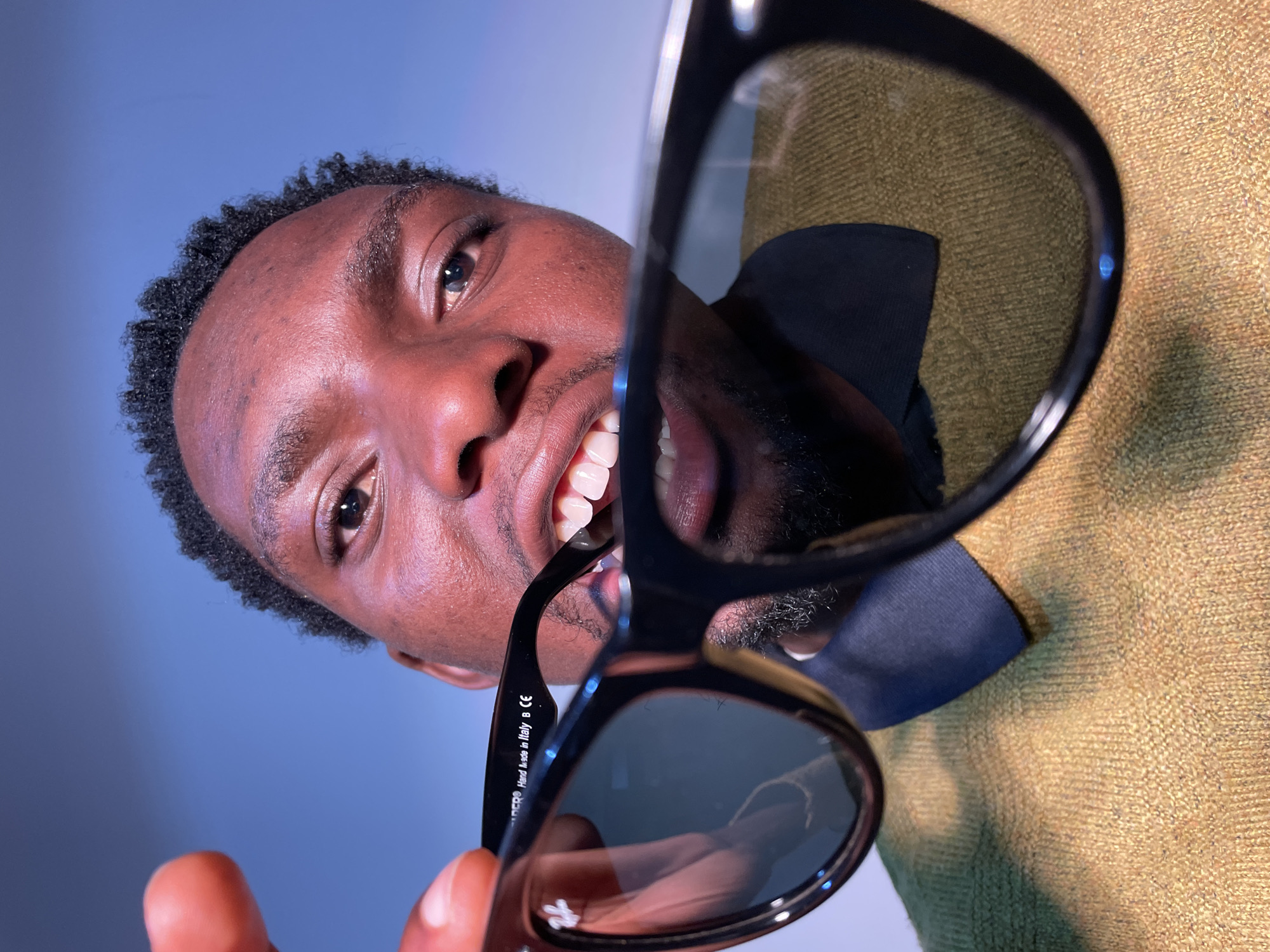 A close-up, low-angle shot of a Black man holding a pair of black sunglasses with a Ray-Ban logo, partially in his mouth. The sunglasses are the main focus, with the man's face slightly blurred behind them. He has short, dark hair and light brown eyes. He is wearing a mustard-yellow sweater with a dark blue collar underneath. The background is a gradient of light blue to white. No information on the city is available in the photo.