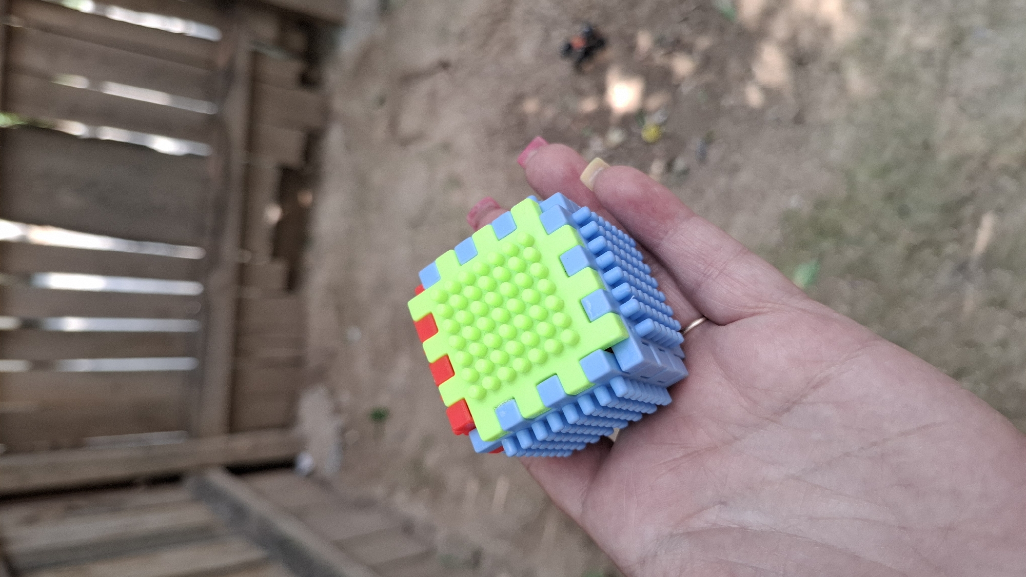 A person's hand, adorned with a ring and painted fingernails in pink and yellow, is holding a small, colorful cube-shaped toy. The toy is constructed from interlocking plastic pieces, predominantly light blue and lime green, with a few red accents on one corner. One face of the cube is entirely covered in small, raised green nubs. The background of the image is blurred, but appears to be an outdoor setting with dry, sandy ground and some vegetation, possibly in Shymkent, Kazakhstan. In the far background, a small, dark object resembling a toy car is visible. The lighting suggests it is daytime, with sunlight filtering through unseen foliage, casting some dappled light and shadows. The overall scene is casual and appears to be a moment of play or a close-up examination of the toy.