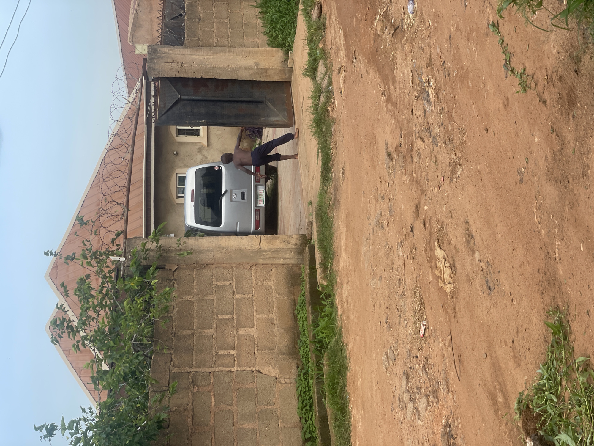 In Bukuru, Nigeria, a young, shirtless boy wearing dark pants stands outside a property, his right arm extended as if pushing open a large, dark metal gate. The gate is slightly ajar, revealing a silver van parked inside the yard. The yard is partially visible through the opening, showing a building with a reddish-brown, corrugated metal roof, adorned with barbed wire. The building has cream-colored walls and at least two windows. A brick wall, roughly constructed and covered in some vegetation, surrounds the property. In front of the wall, a concrete structure with some greenery runs along its base. The foreground depicts a dirt road, uneven and reddish-brown, with scattered debris. The sky above is overcast.
