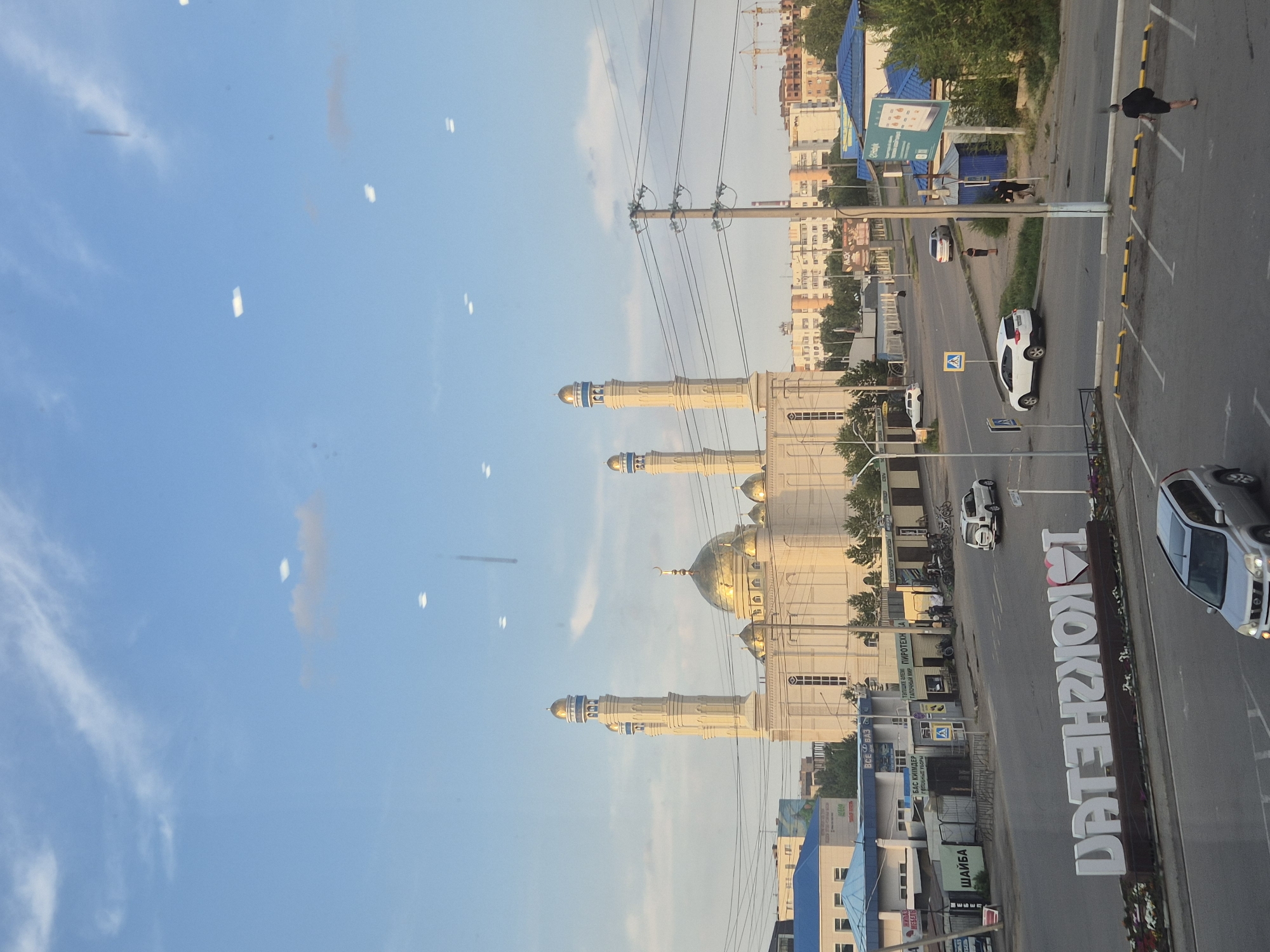 The image captures a daytime view of a city street in Kokshetau, Kazakhstan, focusing on a prominent mosque with golden domes and minarets. The mosque is a central element, flanked by other buildings and overhead power lines. In the foreground, large white letters spelling out "I LOVE KOKSHETAU" are visible, with a heart shape replacing the letter "O." Several cars are on the street, including a silver SUV and a white SUV, and a person is walking in a parking lot. The sky is blue with scattered clouds, and some construction cranes are visible in the distance. The scene appears to be a sunny day with moderate urban activity.
