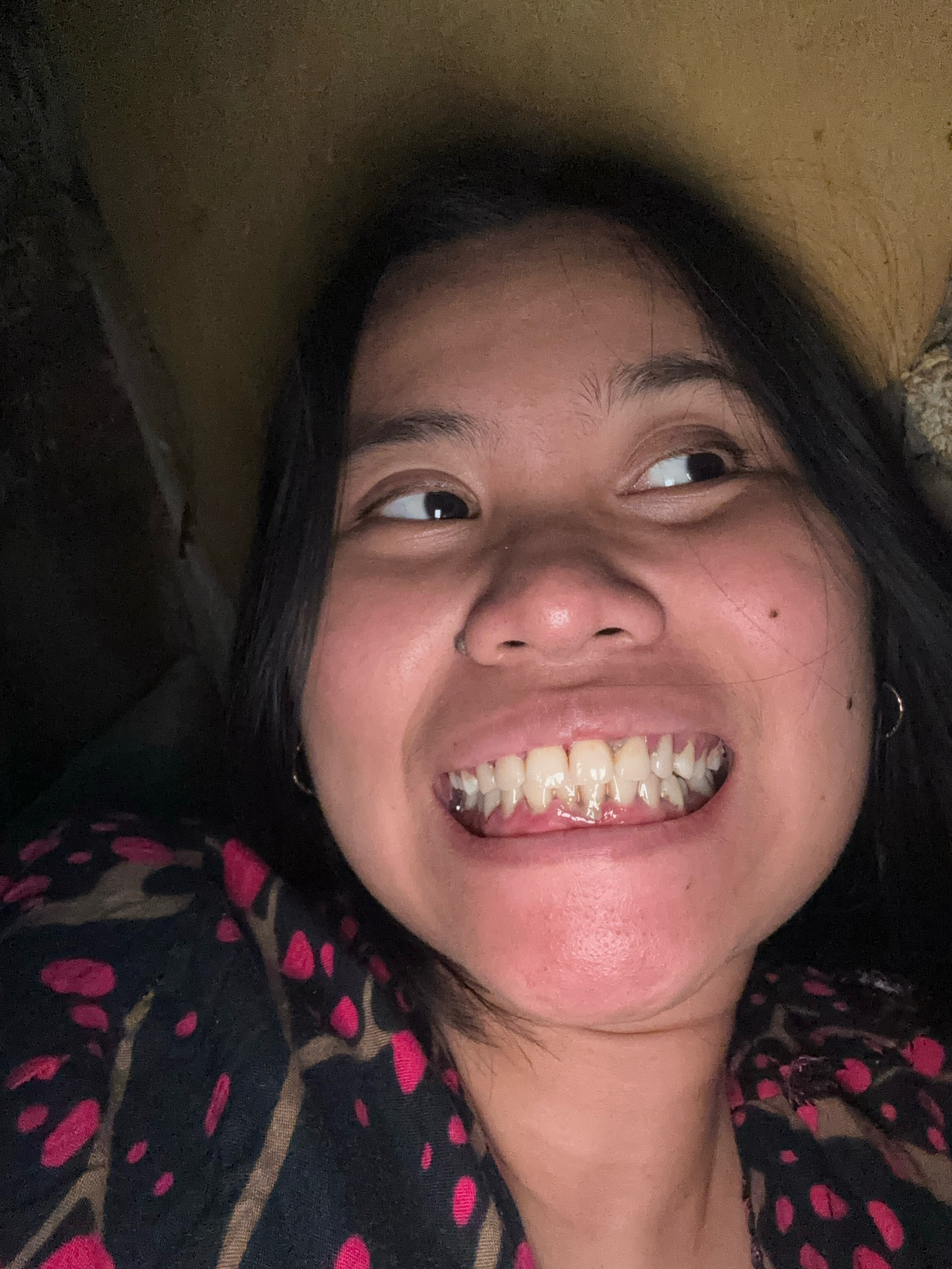 A close-up, selfie-style photograph captures a young woman's face from a slightly low angle. She is looking upwards and to her right, with her mouth wide open in a broad smile, revealing her teeth. Her dark hair frames her face. She is wearing a dark top with a repeating pattern of pink and brown abstract shapes. The background appears to be an indoor environment with a textured, yellowish wall to the upper right and darker, shadowed areas to the left and top left. The lighting suggests an indoor setting, possibly at night. There are no visible landmarks or context to place this image specifically within East Jakarta, Indonesia.