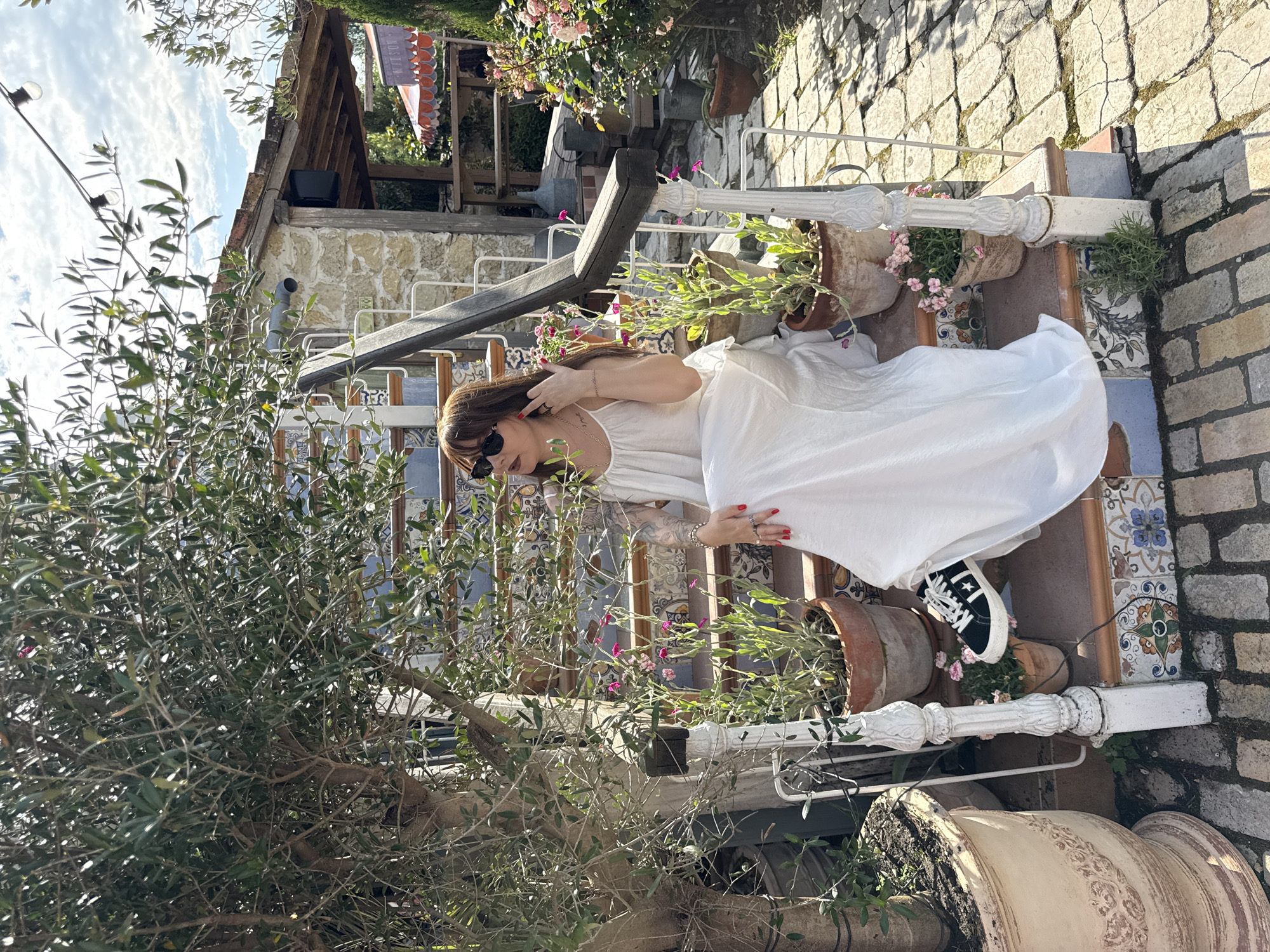 A woman with brown hair and sunglasses is sitting on outdoor stairs. She is wearing a white dress and black Converse sneakers. The stairs are decorated with colorful tiles and lined with potted plants. Behind her, there is a stone building and a large tree with olive branches. The ground is paved with cobblestones. The scene is set in a sunlit outdoor environment, with a blue sky and some clouds visible.