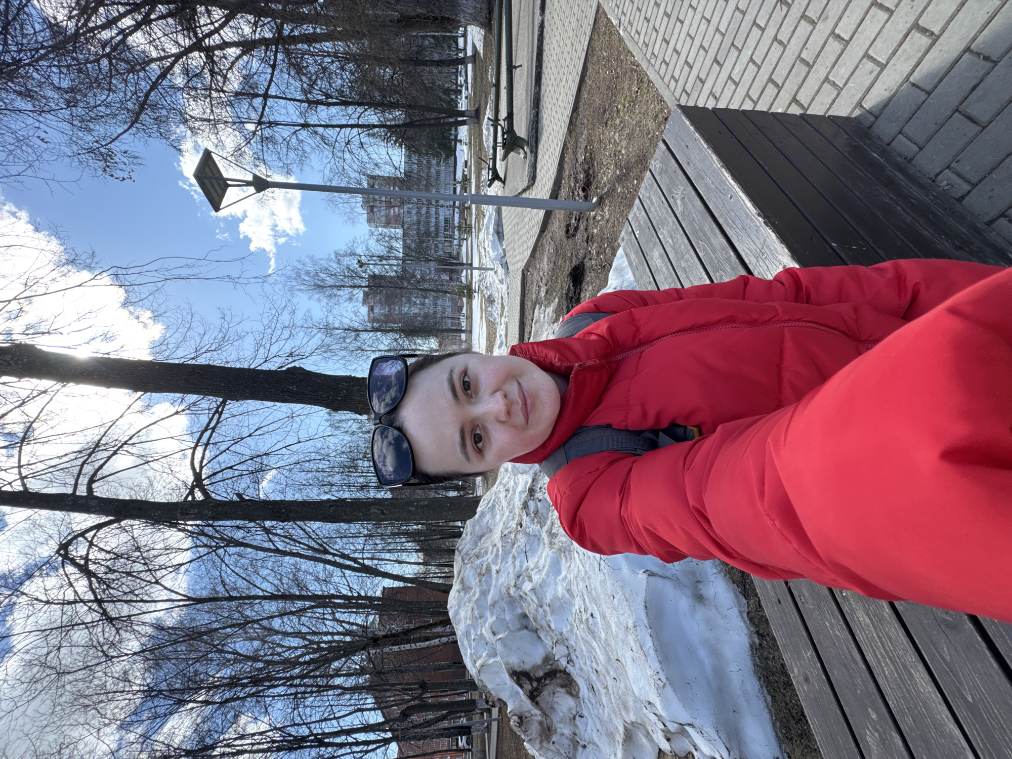 A person wearing a red winter jacket and sunglasses is sitting on a wooden bench in a park. Patches of snow are visible on the ground. Bare trees and a building are in the background. A lamppost stands nearby. The location is possibly in Izhevsk, Russia, though this is not explicitly visible in the image.