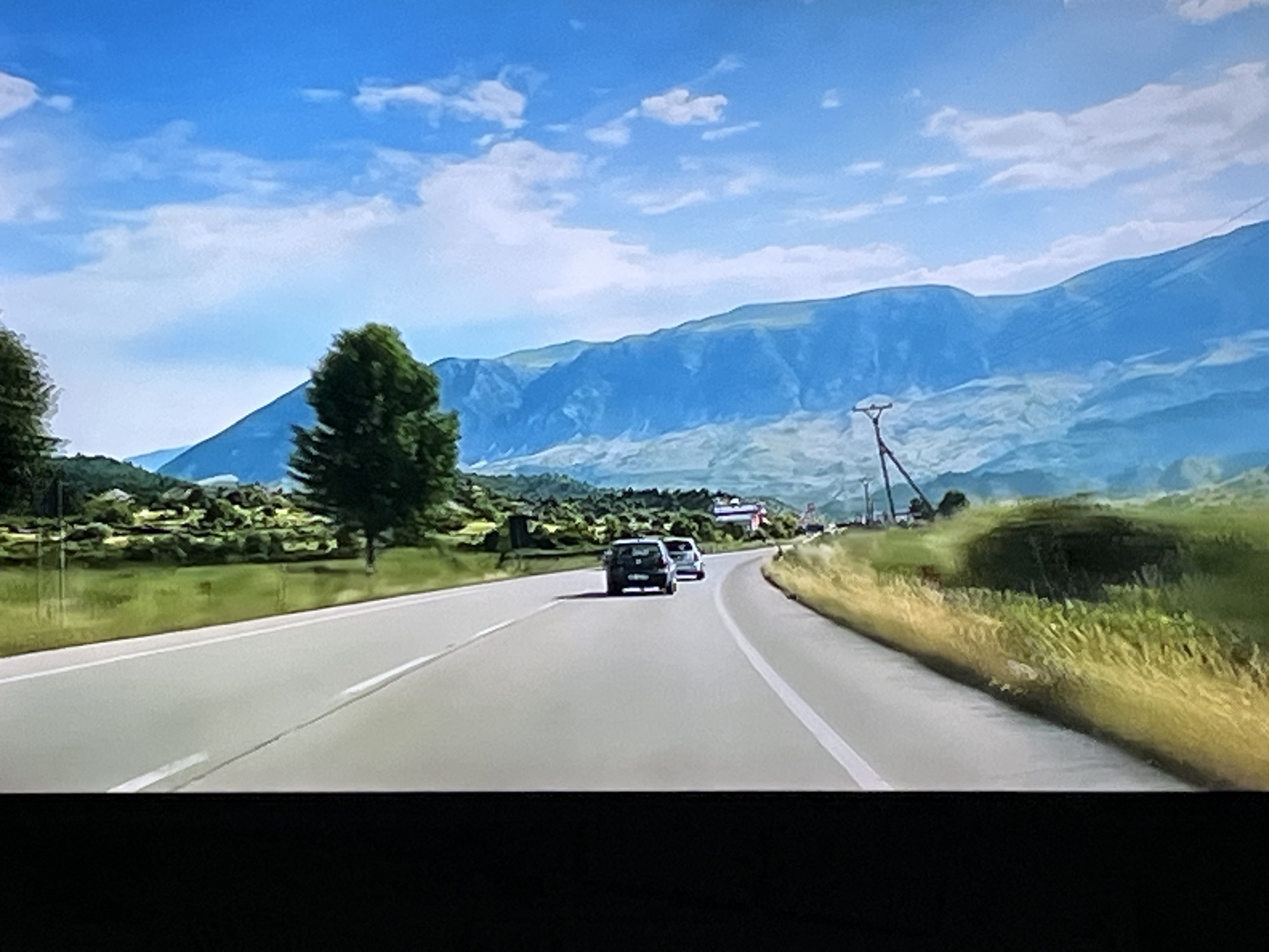 The image depicts a scenic view of a winding road with two cars driving on it. The road is bordered by green grassy fields and trees. In the background, there are mountains under a bright blue sky with scattered clouds. There is no visual information within the image that suggests the location is the city of Kharkiv, Ukraine.