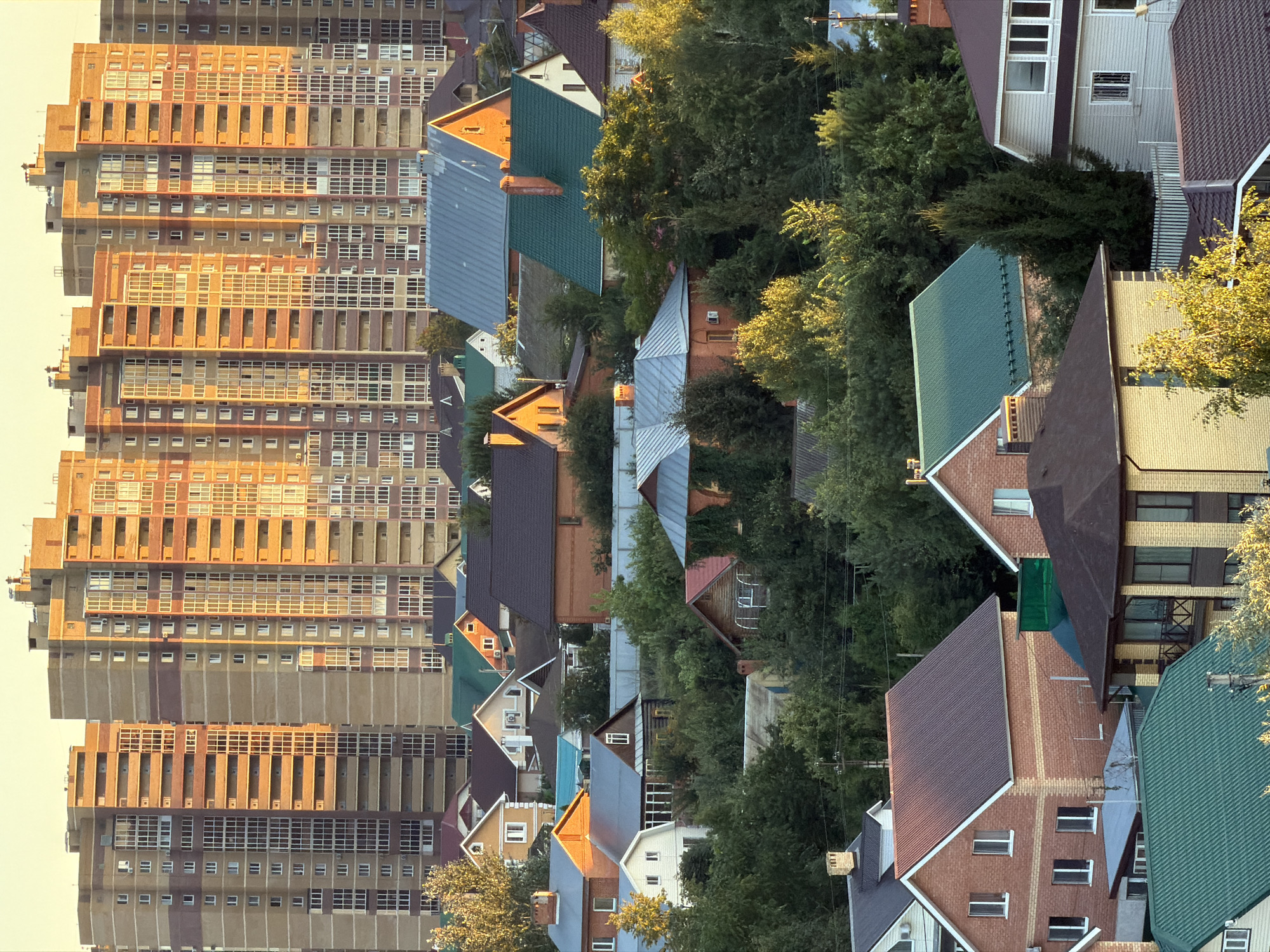 This is an aerial, eye-level, medium shot of a residential area at dusk in Unknown, Unknown. The scene is dominated by a cluster of tall, multi-story apartment buildings in the background, constructed of light brown and reddish-brown brick. Their numerous windows reflect the warm, golden light of the setting sun. In the foreground and middle ground, a dense collection of smaller, individual houses with varying rooflines and colors is visible, interspersed with lush green trees. The houses feature pitched roofs in shades of green, brown, and gray, and are constructed from brick and other building materials. The overall impression is one of a densely populated urban or suburban environment transitioning from day to evening.