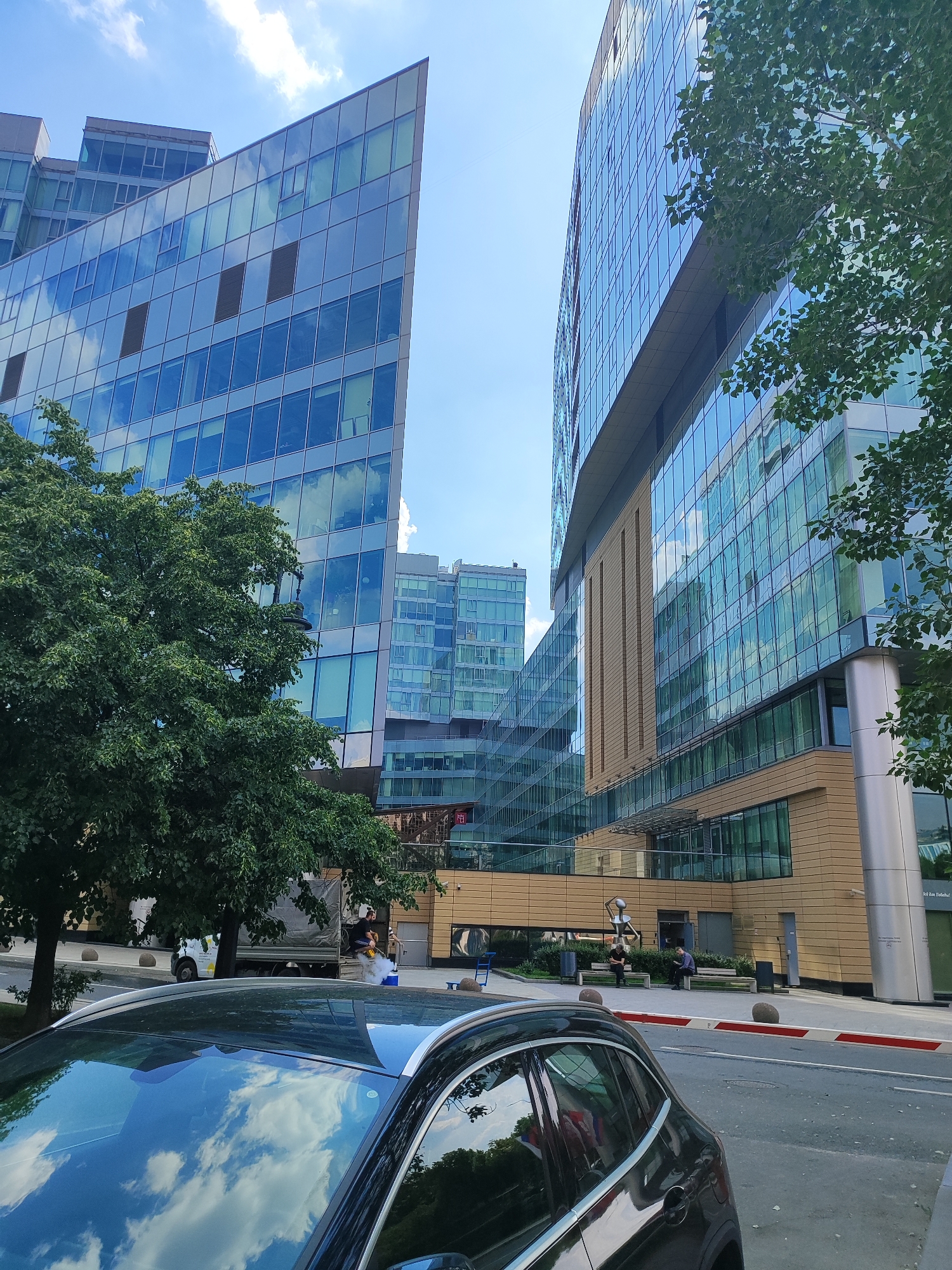 In Moscow, Russia, a modern urban street scene unfolds under a bright, cloudy sky. Two contemporary office buildings dominate the view, their facades clad in reflective blue glass and warm-toned panels. The building on the left features an angular, almost triangular design, while the one on the right is more rectangular, with prominent vertical elements. Green trees line the street, their leaves offering a natural contrast to the architectural structures.
In the foreground, the reflection of the sky and clouds is visible on the polished surface of a dark-colored car. The car's roofline and windshield are prominent. Further down the street, a few individuals are present. Two people are seated on benches outside one of the buildings, one appearing to be engaged with a device. Across the street, a truck and a worker using a steam cleaner are visible, suggesting ongoing street maintenance or cleaning. The area is well-lit, with street lamps and bollards indicating a public space.