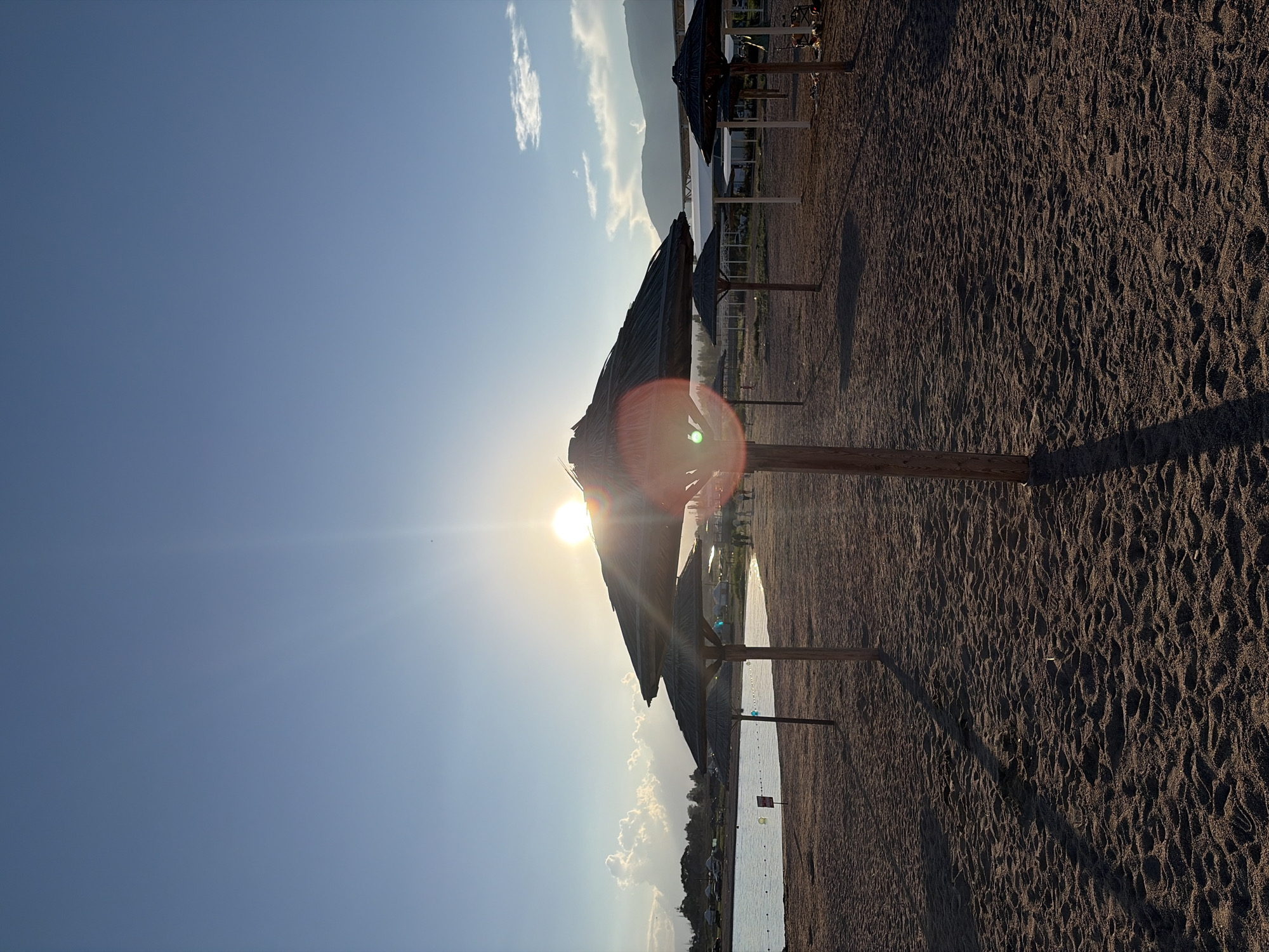 The image captures a serene beach scene at sunset. The foreground is dominated by dark, textured sand, showing ripples and footprints. Several straw umbrellas, supported by wooden poles, are positioned in a row, casting long shadows across the sand. The sun is low on the horizon, appearing as a bright, lens-flared orb behind one of the umbrellas, creating a warm glow and starburst effect. The sky is a clear, deep blue, with a few wispy white clouds scattered near the horizon.
In the middle ground, a calm body of water, likely the sea, is visible. It is bordered by a thin line of vegetation and some distant structures or buildings. A few buoys and a small sign are visible in the water, suggesting a designated swimming or recreational area. The overall atmosphere is peaceful and tranquil, evoking a sense of relaxation. The time of day is clearly sunset, given the low angle of the sun and the warm light. The weather appears to be clear and pleasant. There is no visible text in the image.