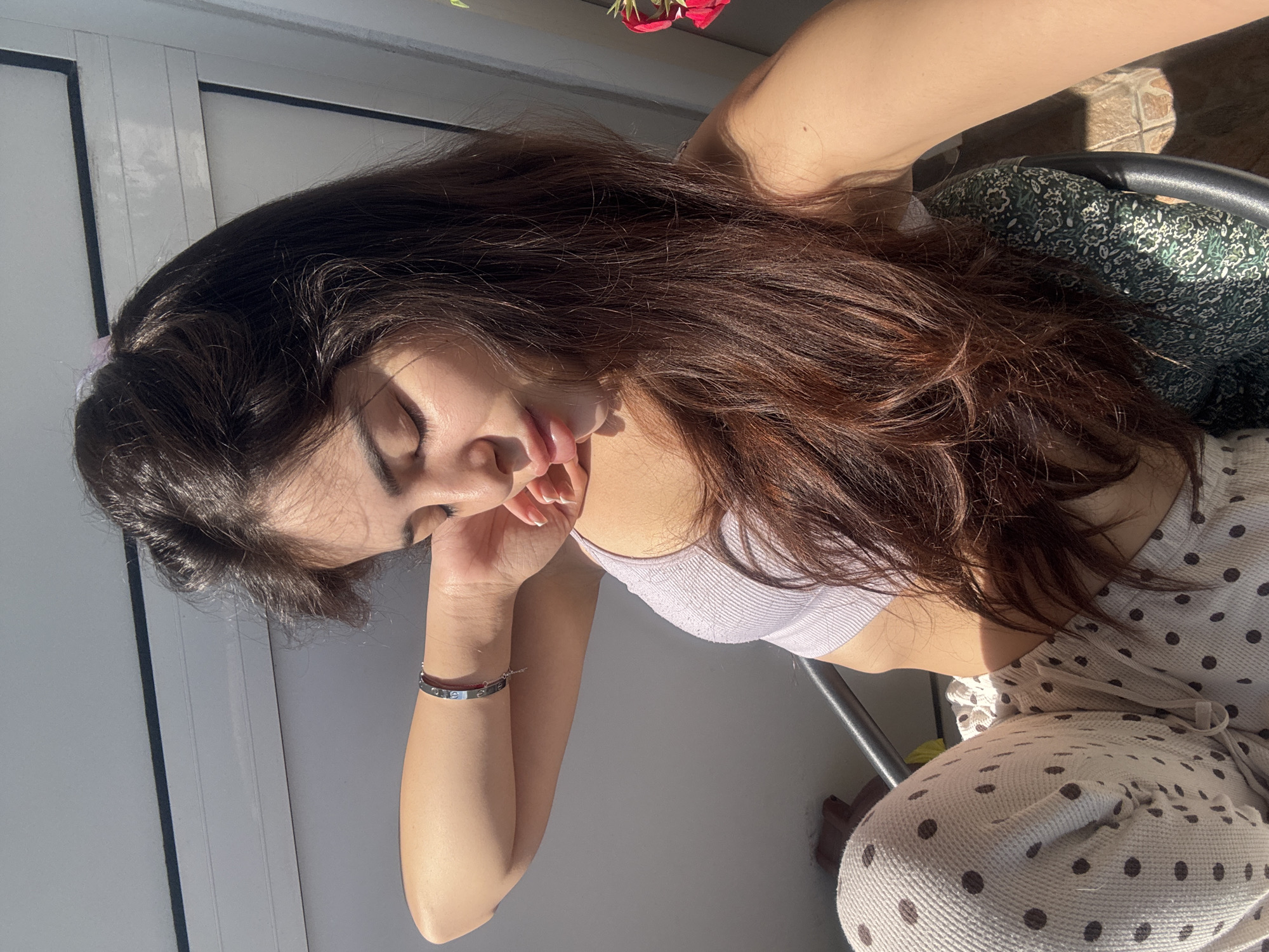 A young woman with long, dark brown hair is captured in a relaxed pose, positioned horizontally in the frame due to the image's rotation. She is shown from the upper torso, with her head resting gently on her left hand, eyes closed or cast downwards.
She is wearing a light lavender or soft purple ribbed bralette or crop top. Her lower body is clad in light-colored (likely off-white or beige) lounge pants or pajamas featuring dark brown polka dots. On her left wrist, a silver-toned bracelet with small red details is visible.
The setting appears to be an indoor space with abundant natural light, possibly near a large window or on a balcony. A light grey wall or window frame provides a clean background. In the upper right corner, a few bright red flowers are visible, suggesting outdoor greenery. A dark chair with a patterned green and white fabric backrest is partially seen behind her. The strong, direct sunlight casts distinct shadows, indicating a bright, sunny day, likely during midday or early afternoon.
The woman's closed eyes and relaxed posture evoke a sense of peace, rest, or enjoyment of the sunlight. No visible text is discernible in the image.