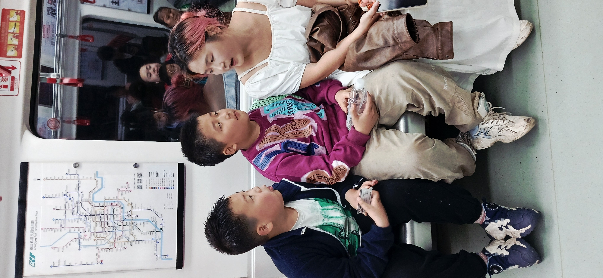 Inside a train car in Unknown, Unknown, two young boys sit on the floor, facing each other. The boy on top wears a purple sweater with a colorful design, tan pants, and white sneakers. He holds a plastic water bottle. The boy on the bottom wears a navy blue jacket over a white t-shirt, black pants, and blue sneakers. He holds a rectangular card. A woman with pink-streaked hair, wearing a white dress, sits on the boy on top with a brown leather jacket to her side and looks at a tablet in her hand. A subway map is visible to the left of the boys. Other passengers are visible through the train's window.