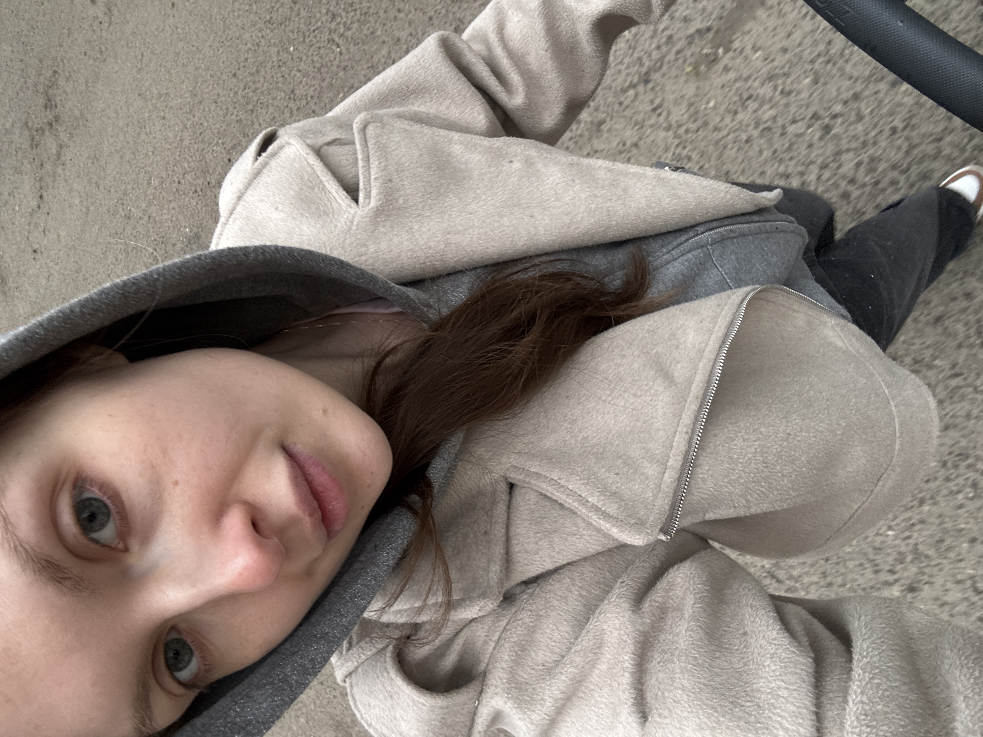 A woman is shown from a high-angle perspective, potentially a selfie. She has fair skin, light-colored eyes, and light brown hair. She is wearing a gray hooded sweatshirt and a light beige fleece jacket. Below the jacket, a gray fabric is visible. She is wearing dark pants and one visible white sneaker with a brown stripe. The background appears to be a gray concrete surface. The location is unspecified; there is no indication that it is in Moscow, Russia.