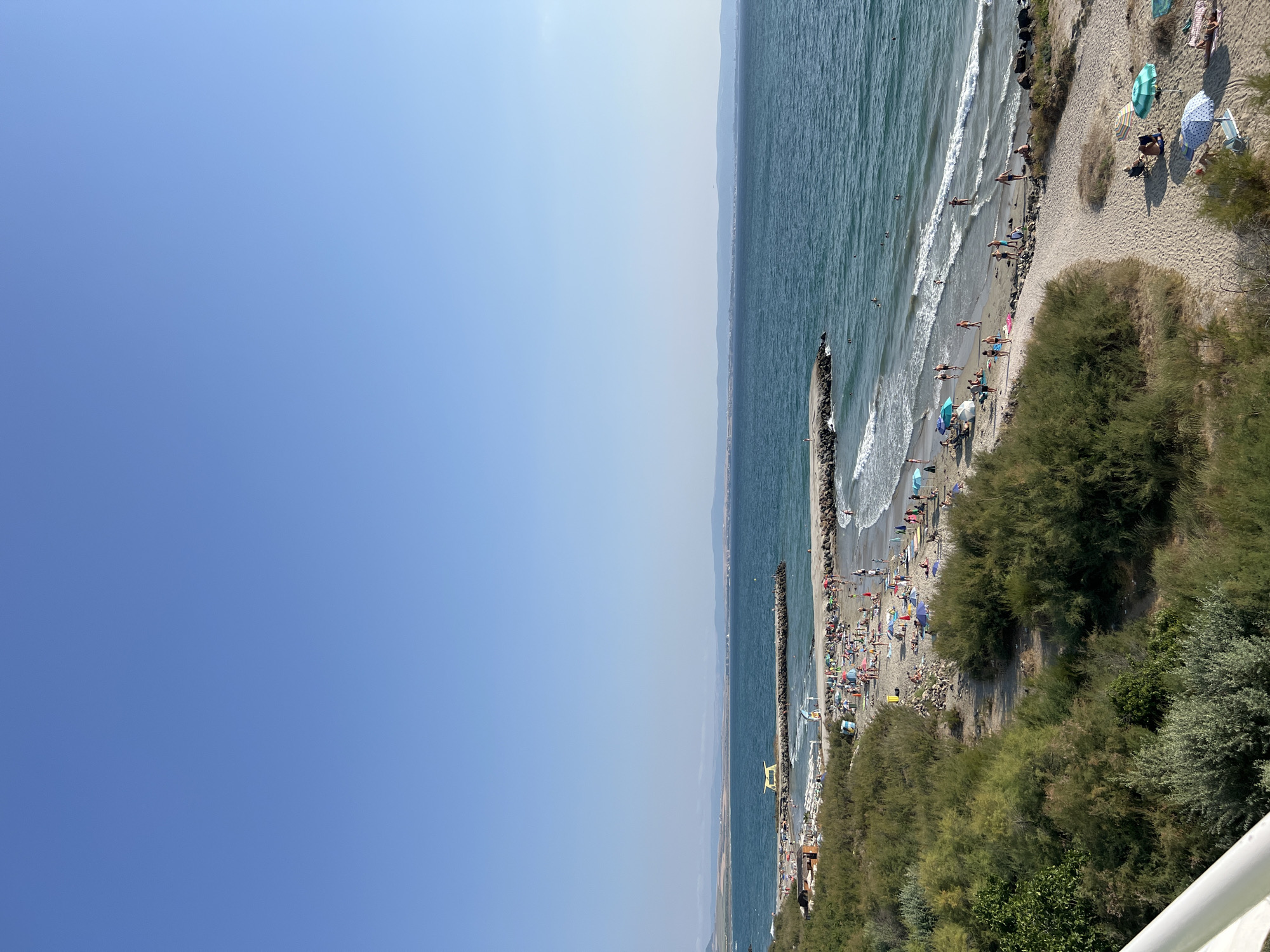This image depicts a sunny day at the beach in Pomorie, Bulgaria. The scene is a wide shot of a bustling beach with many people enjoying the water and sand. In the foreground, there is a stretch of dry grass and bushes bordering the sandy beach. Moving towards the water, the sand is covered with beach umbrellas and towels, with several individuals relaxing.
The sea is a vibrant blue, with moderate waves reaching the shore. A stone pier extends into the water, featuring a yellow structure on its end, possibly a lifeguard station or observation post. Along the beach, numerous people are seen swimming, walking, sunbathing, and playing in the water. The background shows a distant coastline with buildings and hills under a clear blue sky. The overall atmosphere is one of a lively summer day at a popular seaside resort.