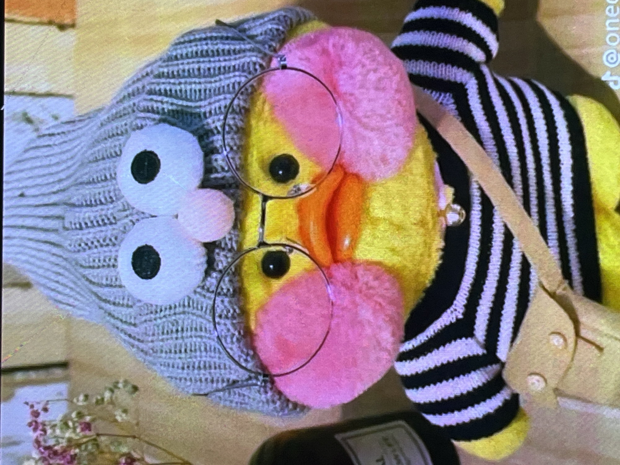 This media file features a close-up, slightly blurry shot of a stuffed toy resembling a duck. The duck is wearing a knitted gray sweater, a black and white striped shirt, and a small beige bag slung across its body. It has large, white circular eyes with black pupils and pink fluffy cheeks. A pair of thin, round glasses is perched on its beak. The background is a warm-toned, textured surface, possibly wood, with some out-of-focus pink and white flowers in the lower left corner and a dark bottle in the foreground. The lighting appears to be artificial, casting soft shadows. There is no specific activity taking place; the toy is posed. The overall impression is whimsical and cute. A faint watermark with the text "@onec" is visible in the upper right corner. The location context provided is Cadek, Indonesia.