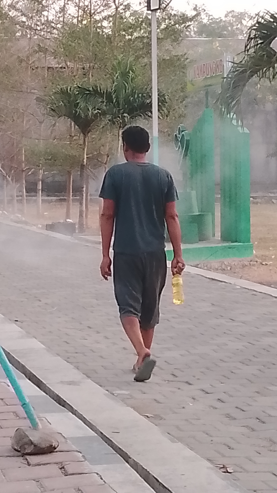 The image captures a man walking away from the camera on a paved pathway in what appears to be a public outdoor space, likely in Karanglangu, Indonesia. The man is wearing a dark t-shirt and dark shorts, and he is carrying a yellow plastic bottle in his right hand. His left hand is holding a lit cigarette. He is wearing open-toed sandals. In the background, there are trees, a lamppost, and green structures that might be part of a recreational area or a building. A sign with the word "KAMPUNGKB" is visible in the upper right background, indicating the location. A light mist or haze is present in the air, particularly around the green structures. The pathway is made of interlocking pavers, and a concrete curb runs along the left side of the path. A turquoise pole with a stone base is also visible on the left side of the frame, near the curb. The lighting suggests it is either early morning or late afternoon.