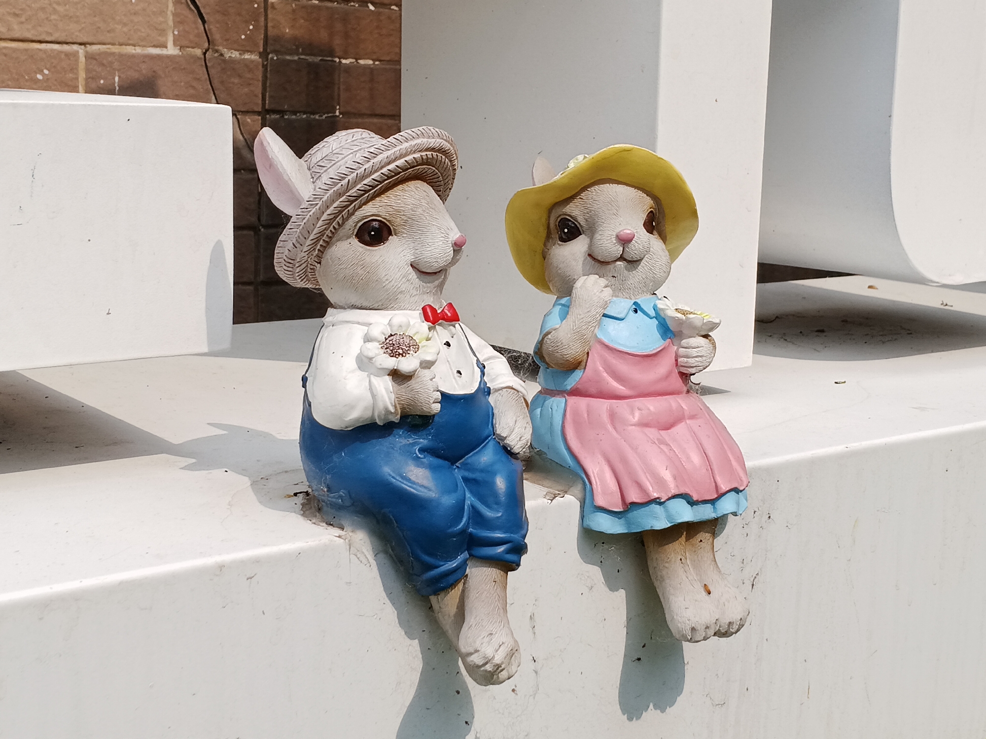 Two charming rabbit statues are prominently featured, sitting side-by-side on a white ledge under bright, direct sunlight. The statue on the left, depicted as a male rabbit, wears a textured straw hat, a white long-sleeved shirt, blue overalls, and a small red bow tie. He holds a white daisy-like flower with a yellow center in his left paw. His companion, a female rabbit on the right, sports a yellow hat, a light blue top, and a pink apron or dress. She holds a small, light-colored object, possibly a tiny flower or berry, in her paw. Both rabbits have smiling expressions, pink noses, and large, dark eyes, conveying a cheerful and friendly demeanor. Their legs dangle over the edge of the white surface they are seated on.
The statues appear to be made of painted ceramic or resin, showing some minor signs of outdoor exposure such as dust and slight discoloration, particularly on their feet and the base. Strong shadows are cast by the statues and the white structures, indicating a clear, sunny day. In the background, a dark, weathered brick wall with visible cracks and stains provides a contrasting backdrop to the bright white elements. The overall setting suggests an outdoor decorative display, possibly in a garden, park, or public area within Shunde District, China. There is no visible text in the image.
