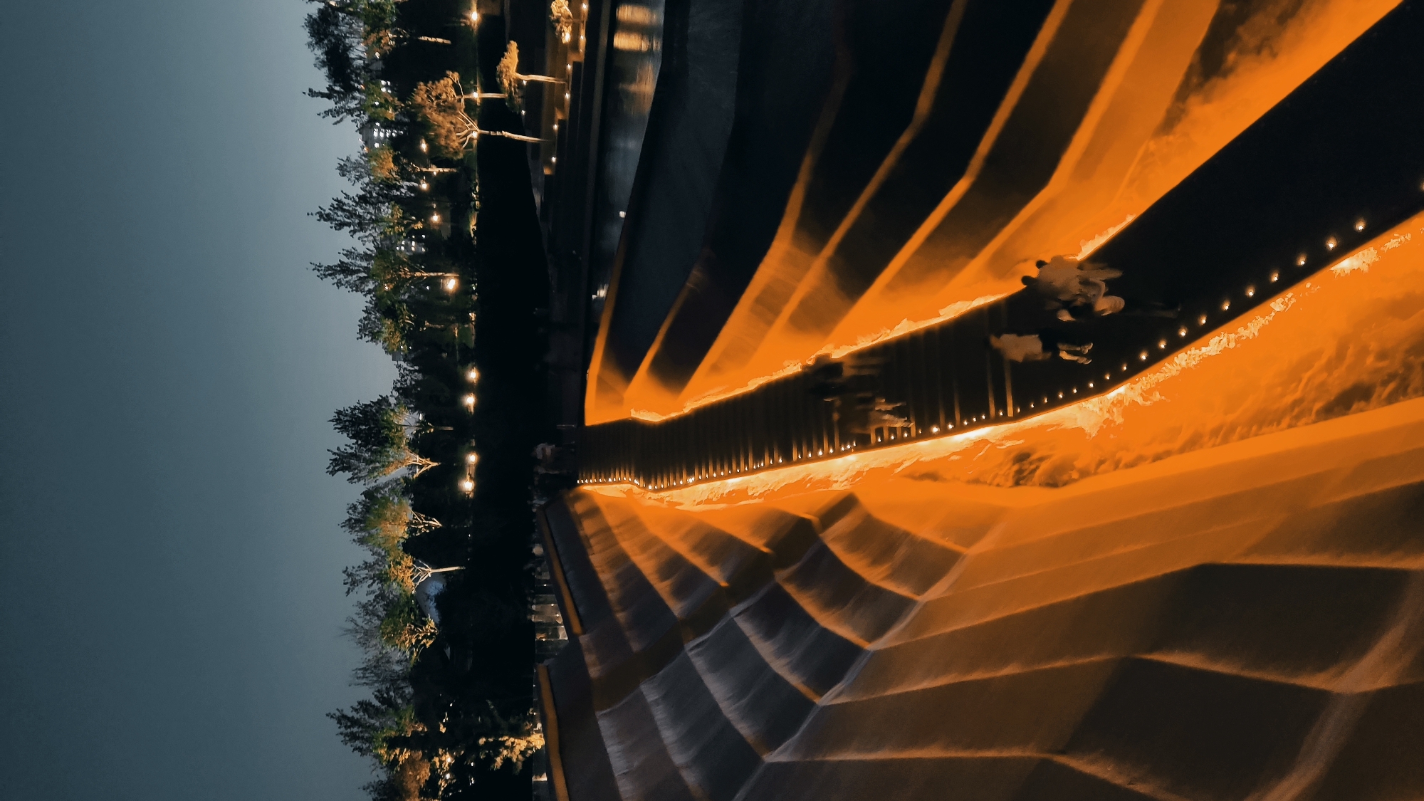 The image depicts a night scene at the "Fountains of Europe" park in Krasnodar, Russia. A tiered water cascade structure dominates the foreground, illuminated with warm orange light along its edges and steps. A pathway, also lined with lights, runs along the center of the cascade, where several individuals are walking. Beyond the cascade, a dark body of water reflects the lights of trees and distant buildings. The sky is a deep twilight blue, with scattered lights from the city visible on the horizon. The overall impression is one of a well-lit, modern urban park at night.