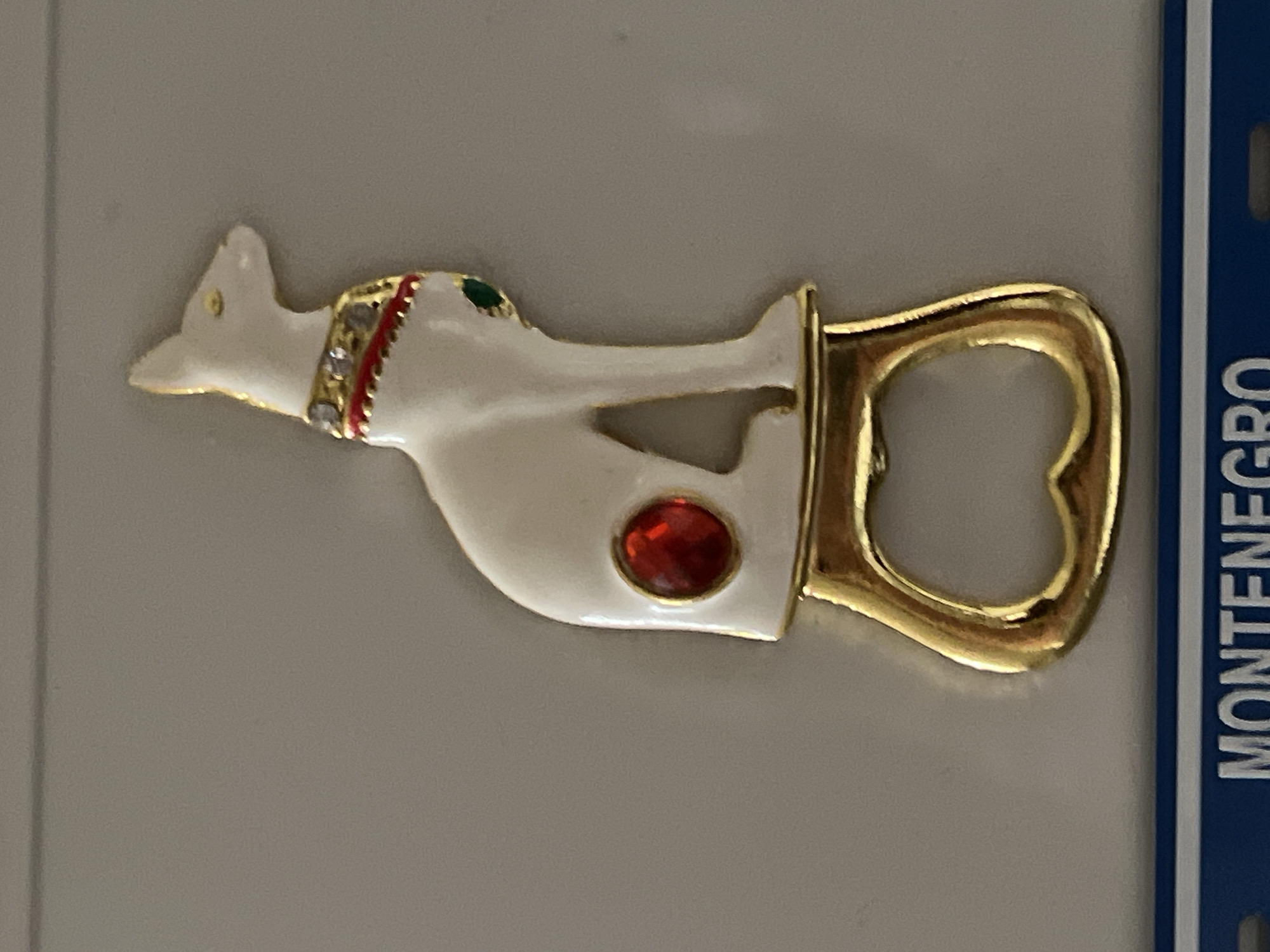 The image shows a white, cat-shaped bottle opener with gold trim. The cat figure has a red jewel on its body, and a jeweled collar. The bottle opener is lying on a gray surface next to a blue strip with the word "MONTENEGRO" vertically printed on it. There is no evidence suggesting the image was taken in Yerevan, Armenia.