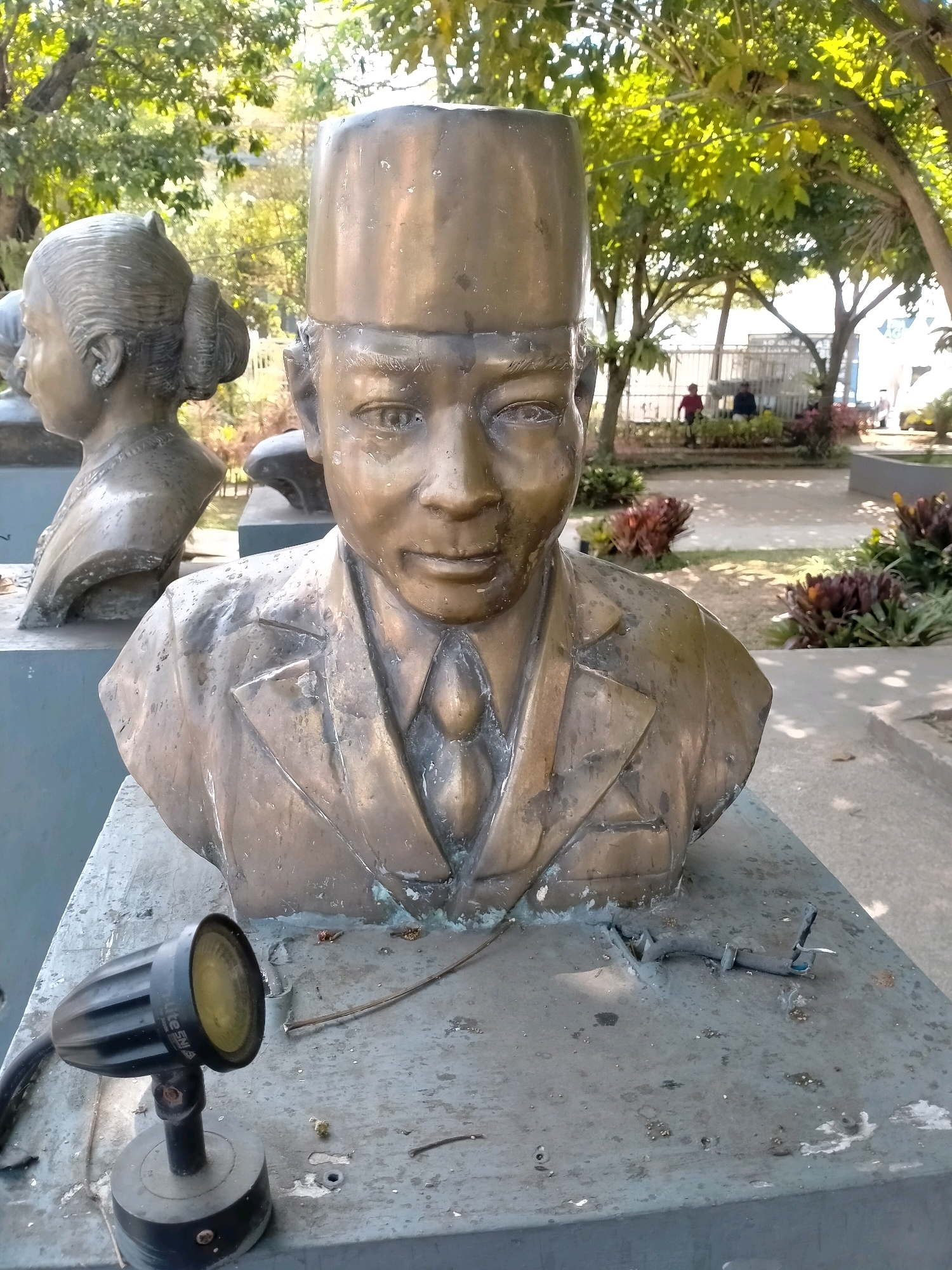 This is a photograph of two bronze busts on a pedestal, located in a park in the city of Batu, Indonesia. The bust in the foreground is of a man wearing a hat and a suit with a tie. The bust behind and to the left is of a woman with her hair styled in a bun. Both busts are weathered and show signs of age. In front of the man's bust, on the pedestal, is a black spotlight. The background of the image shows trees with green leaves and a fence with people walking in the distance. The overall scene appears to be an outdoor public space.