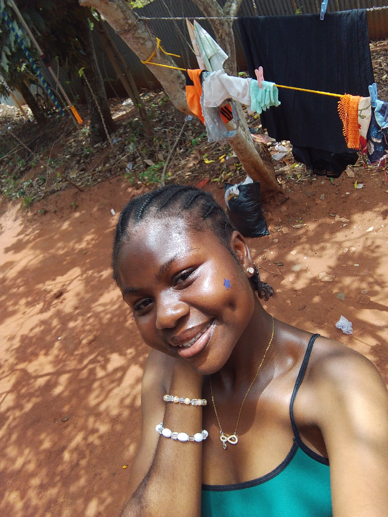 A smiling woman with braided hair is the central focus of the image. She is wearing a green and black tank top, a gold necklace with an infinity pendant, and beaded bracelets. A blue star sticker is on her left cheek. Clothing is hanging on a clothesline in the background, strung between two trees. The ground is reddish-brown and covered in leaves and shadows. A black plastic bag sits on the ground near the base of one of the trees. The location appears to be an outdoor area, possibly a residential yard or a shaded area on the outskirts of the city of Nsukka, Nigeria.