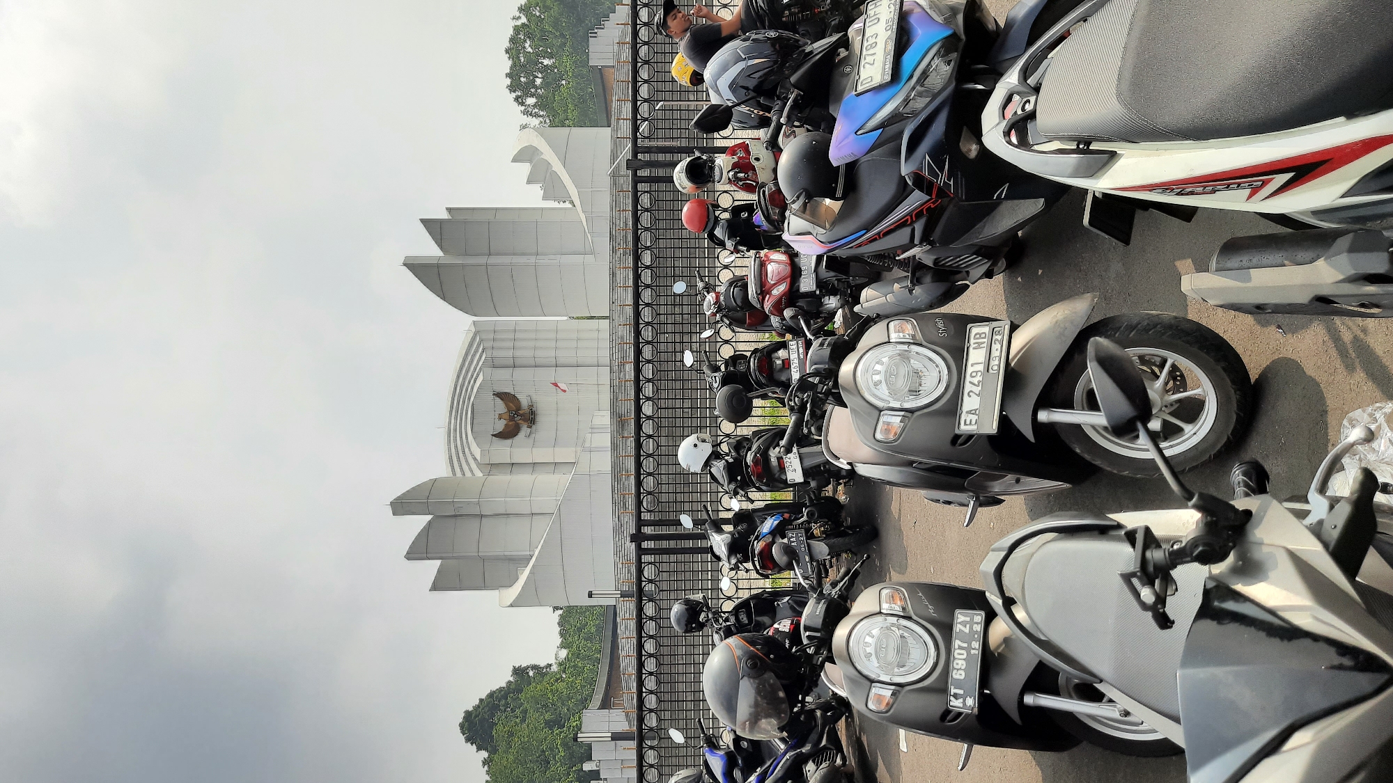 A wide-angle, eye-level shot captures a parking lot filled with a variety of motorcycles, predominantly scooters, in front of a large, modern building. The building, likely a government or civic institution, features a distinctive white facade with geometric, tiered structures and a prominent emblem of a bird of prey, possibly the Garuda Pancasila, centered above an arched entrance. A small Indonesian flag flies to the right of the emblem. The sky is overcast and a muted gray, suggesting either dawn, dusk, or a cloudy day. Lush green trees flank the building on the left side.
The foreground is dominated by parked motorcycles. Many have helmets placed on their seats or handlebars. The motorcycles are of various colors, including black, dark gray, red, blue, and white. Some have visible license plates, such as "KT 6907 ZY" and "EA 2491 NB". The ground is asphalt.
In the mid-ground, behind the motorcycles, is a black metal fence. Beyond the fence is a paved area with stairs leading up to the building. A few people are visible in the distance, near the fence and the building entrance. One person in the far right of the mid-ground is wearing a black shirt and a dark cap and appears to be smoking.
The overall impression is a public space, likely a monument or government building in a city like Bandung, Indonesia, given the architectural style and the presence of the Garuda emblem, which is the national symbol of Indonesia. The motorcycles suggest a common mode of transport for visitors or staff.