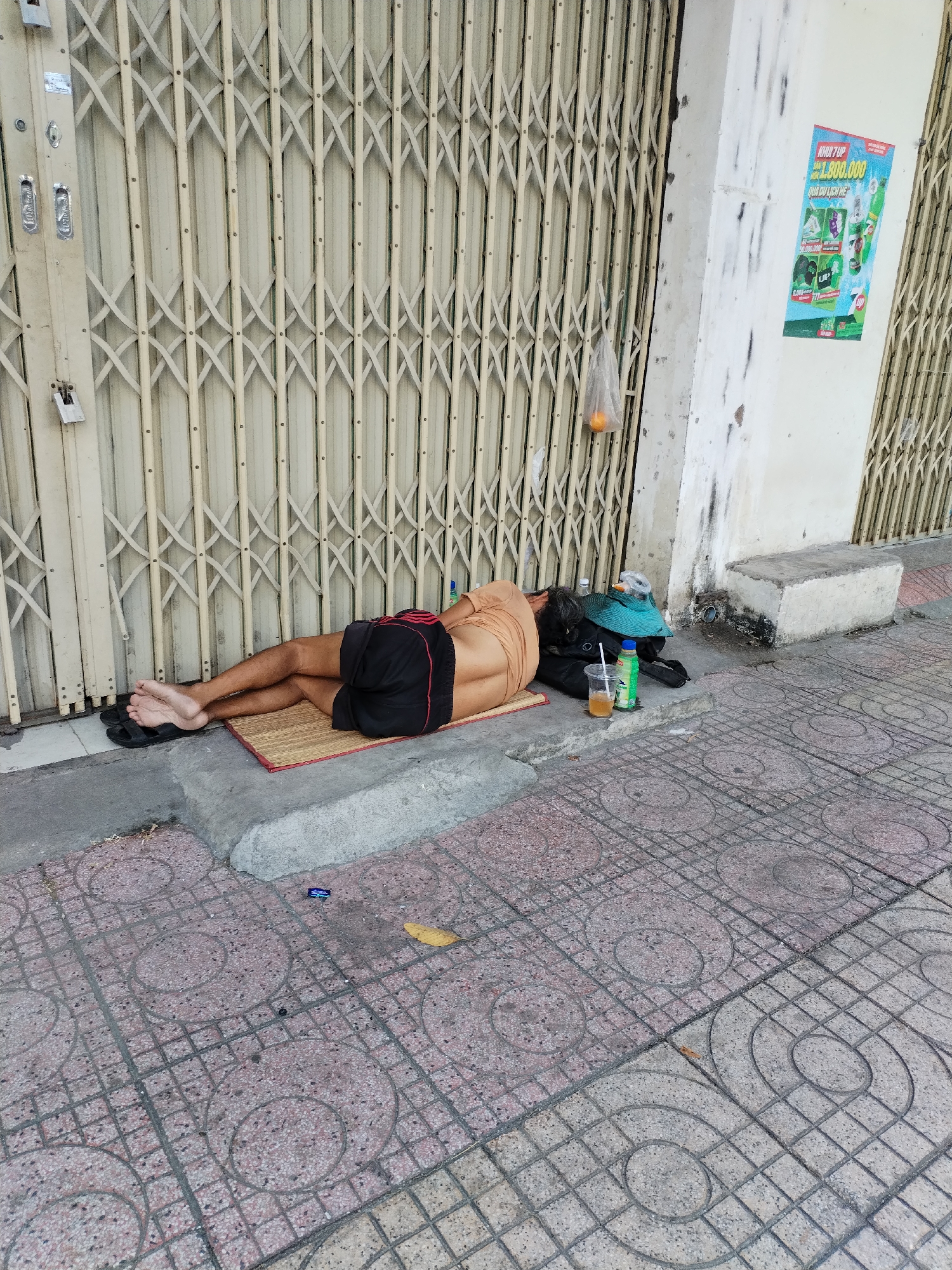 A man sleeps on a woven mat on a sidewalk in Nha Trang Ward, Vietnam. He lies next to a closed accordion-style security gate. The man wears black shorts with red stripes, a tan shirt pulled up to expose his back, and black sandals. A green woven hat, a black bag, and a beverage container are beside him. A plastic bag with an orange hangs from the gate. An advertisement is fixed on the nearby wall. The sidewalk is patterned with circular designs.