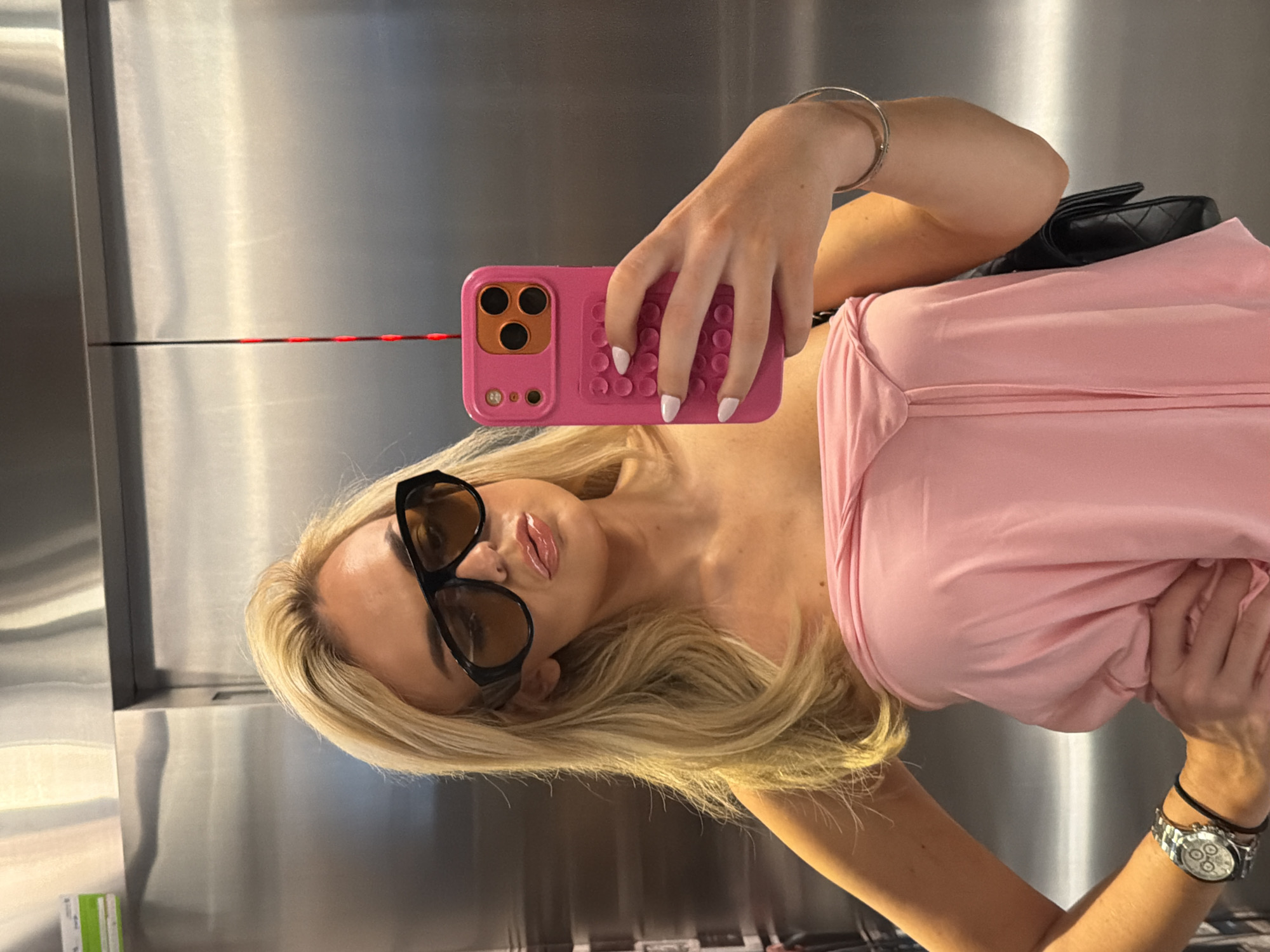 The image depicts a woman taking a selfie inside an elevator in Barcelona, Spain. The elevator interior features brushed stainless steel panels, with a horizontal seam visible across the center. A thin black cord with small red segments hangs vertically from the ceiling in the background, to the right of the woman's head. On the upper left panel, a small rectangular green and white sticker is affixed.
The subject is a woman with long blonde hair, styled around her shoulders. She is wearing large, dark-framed sunglasses with amber-tinted lenses. Her lips are glossy. She wears a strapless, light pink top made of a soft, flowing fabric. Her left hand rests on her hip, holding down the side of her top, and she wears a silver watch with a white dial and multiple subdials on her left wrist, along with a black band. Her right hand holds a bright pink smartphone, which she is using to capture the image. The phone case has a raised, grid-like pattern on its back, resembling push-pop bubbles. The phone's camera module, featuring three lenses, is highlighted by an orange background within the pink case. Her fingernails are painted white. A black quilted handbag is visible behind her left arm.