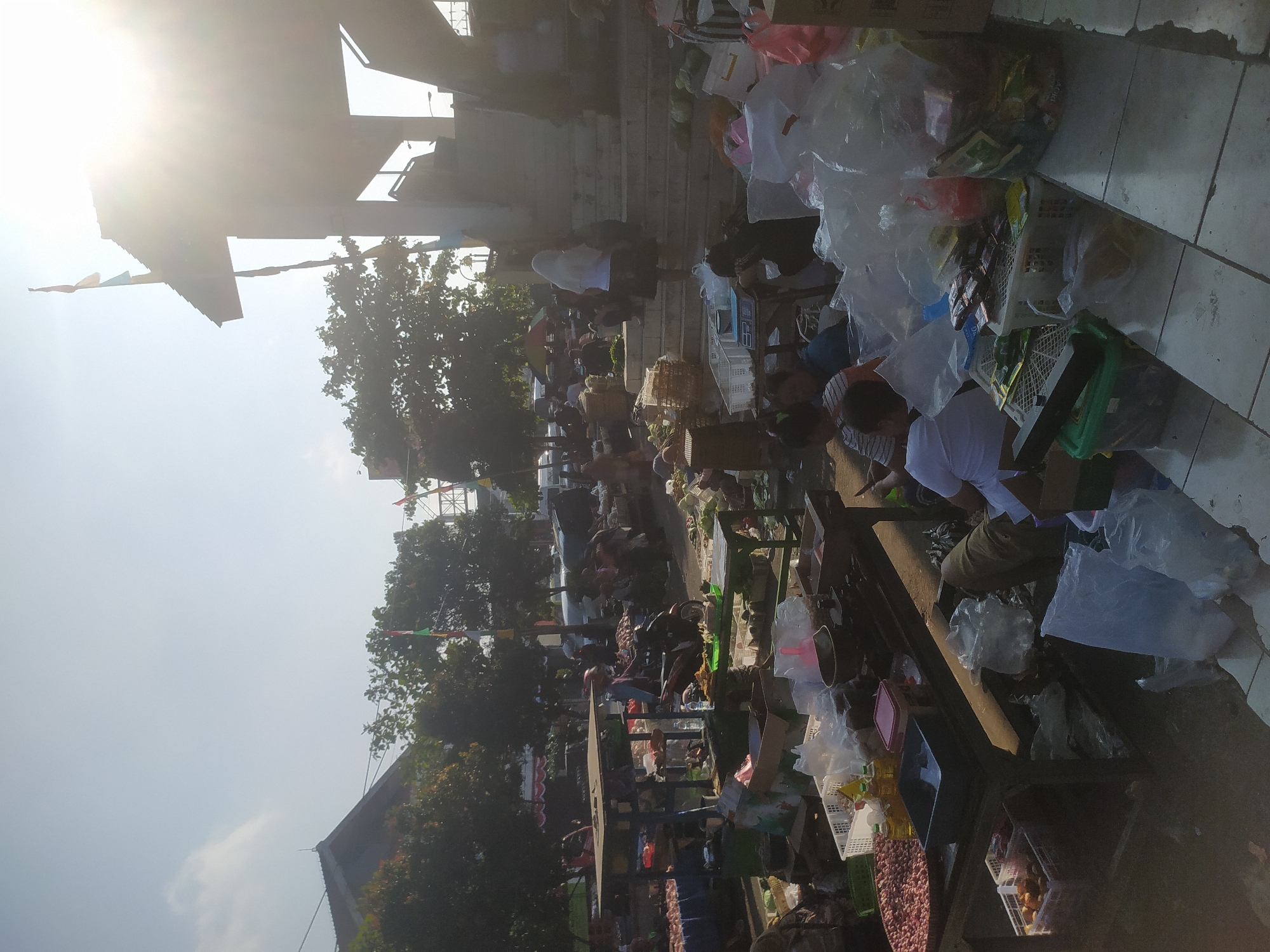 The image depicts a bustling outdoor market scene in Kupang, Indonesia, under a bright, sunny sky with visible lens flare. The market appears to be set up on paved ground, with various stalls and makeshift tables laden with goods.
In the foreground, to the left, is a long, dark wooden table displaying a large quantity of red onions in a round, shallow container. Adjacent to it are bottles of cooking oil and a dark blue plastic container. Further to the right, other items are visible, including baskets of produce and various packages wrapped in plastic.
Several people are present throughout the scene, engaged in activities typical of a market. Some are standing behind stalls, attending to customers or organizing merchandise. Others are seated or kneeling, interacting with goods or resting. A woman in a white headscarf and dark skirt is seen walking up a set of stairs on the right side of the image, towards a covered structure.
The environment includes several trees scattered throughout the market area, providing some shade. In the background, there are buildings, some appearing to be part of the market structure with a covered roof. Flags are visible, possibly indicating a national holiday or local celebration. A motorcycle is parked amongst the stalls, suggesting transportation within the market. The overall atmosphere is one of lively commerce.