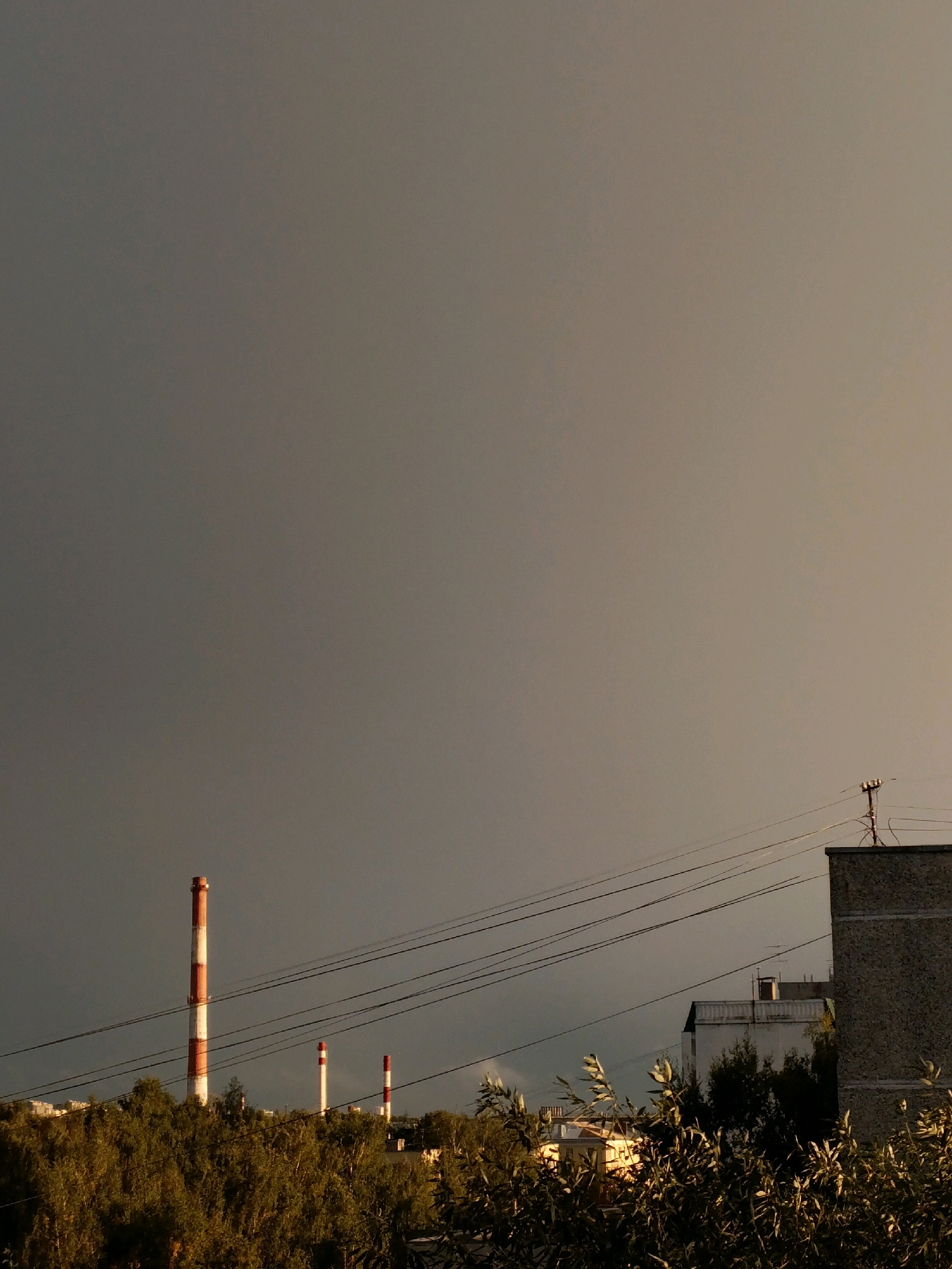 The image captures an outdoor scene featuring industrial chimneys and residential buildings under a cloudy sky. In the foreground, a dense cluster of trees with green and golden foliage is visible. Behind the trees, several power lines stretch across the frame, supported by poles.
Dominating the middle ground are three tall industrial chimneys, two of which are prominently striped in red and white. The largest chimney stands to the left, while two smaller ones are positioned further to the right.
In the background, residential buildings are partially visible. A large, grey, concrete building with a flat roof is on the right side of the frame, with a small structure on its roof that appears to be an antenna or communication equipment. To the left of this, a lighter-colored building with a balcony and a smaller structure on its roof is also present. Further back, some lighter-colored, multi-story buildings with windows can be seen, suggesting a residential area.
The sky is overcast with a dark grey hue, but a soft, warm light on the right side suggests the presence of sunlight breaking through or reflecting off the clouds. The overall lighting creates a somewhat dramatic and moody atmosphere. There are no people visible in the image. The scene appears to be set in an urban or semi-urban environment with a mix of industrial and residential elements.