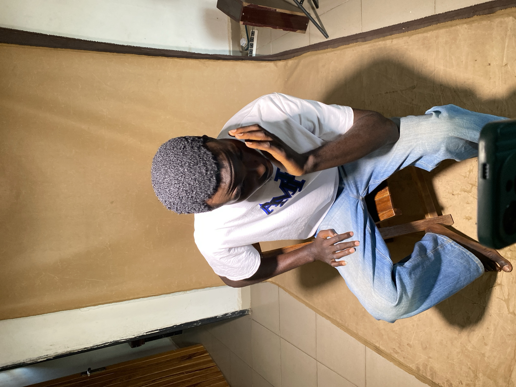 A man wearing a white t-shirt and jeans sits on a wooden chair in a room with beige walls and a white baseboard. He has short, textured hair and dark skin. He is looking down and appears to be touching his face with his left hand. A portion of a black object, possibly a camera, is visible on the right side of the frame. The room appears to be in a residential or office building located in Jos, Nigeria, based on cultural cues and local knowledge.